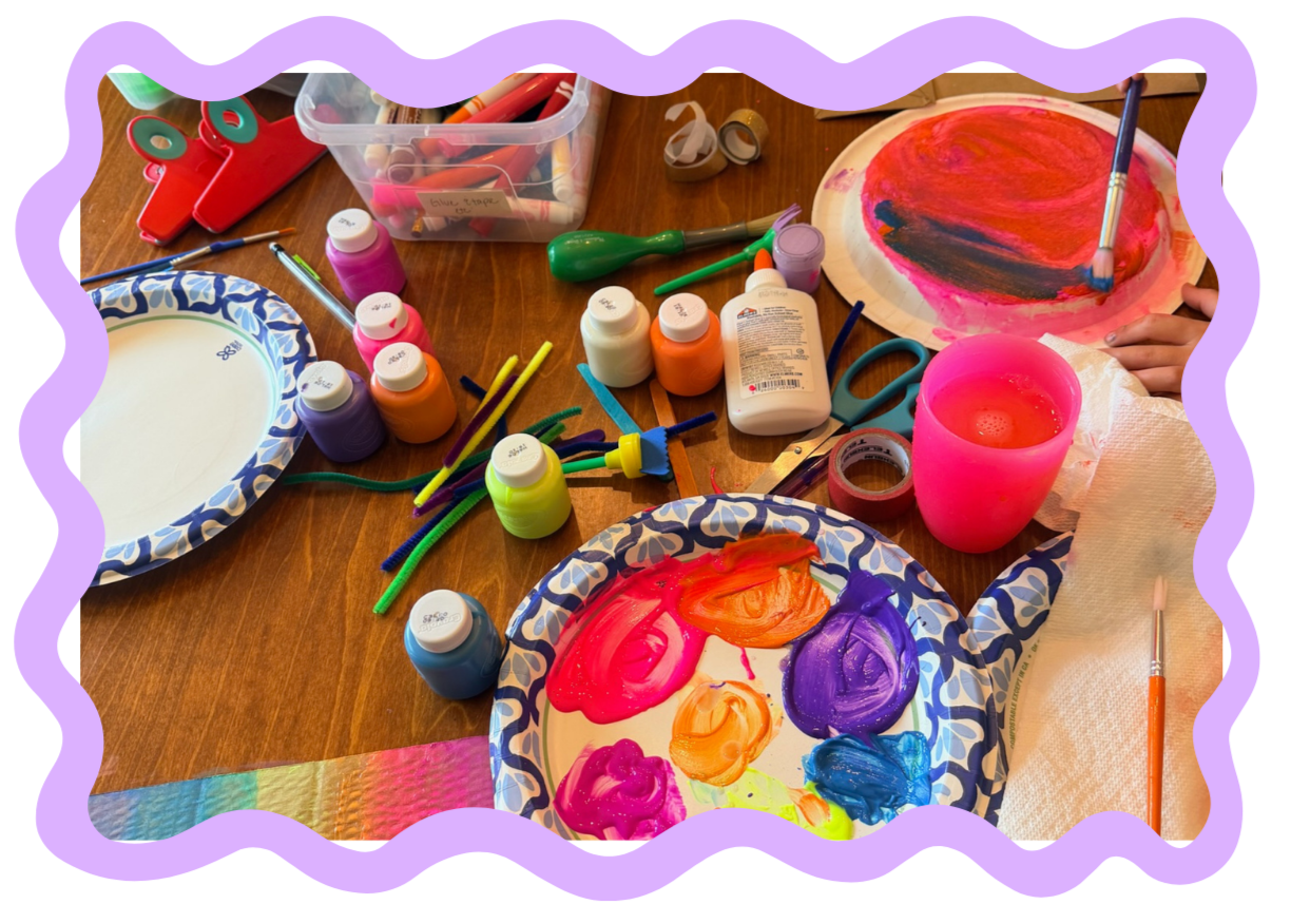 Table with arts and crafts supplies, including paint bottles, paper plates with paint, brushes, scissors, washi tape, and pipe cleaners, as children create colorful painted artwork.