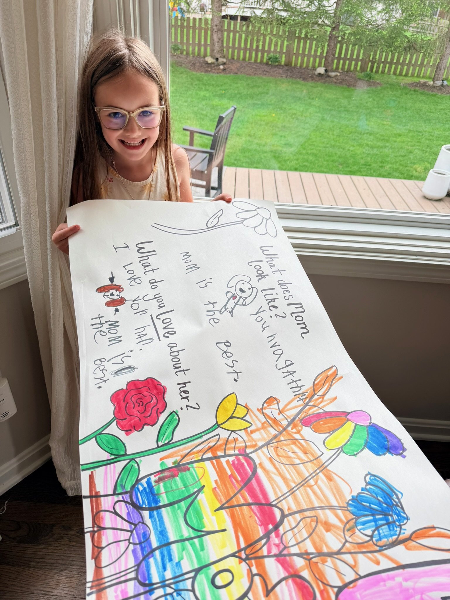 A young girl with glasses and long hair is standing by a window, holding a large colorful poster with handwritten text and illustrations of flowers, butterflies, and rainbow colors. The outdoor background shows a green lawn, trees, a wooden fence, and a patio.