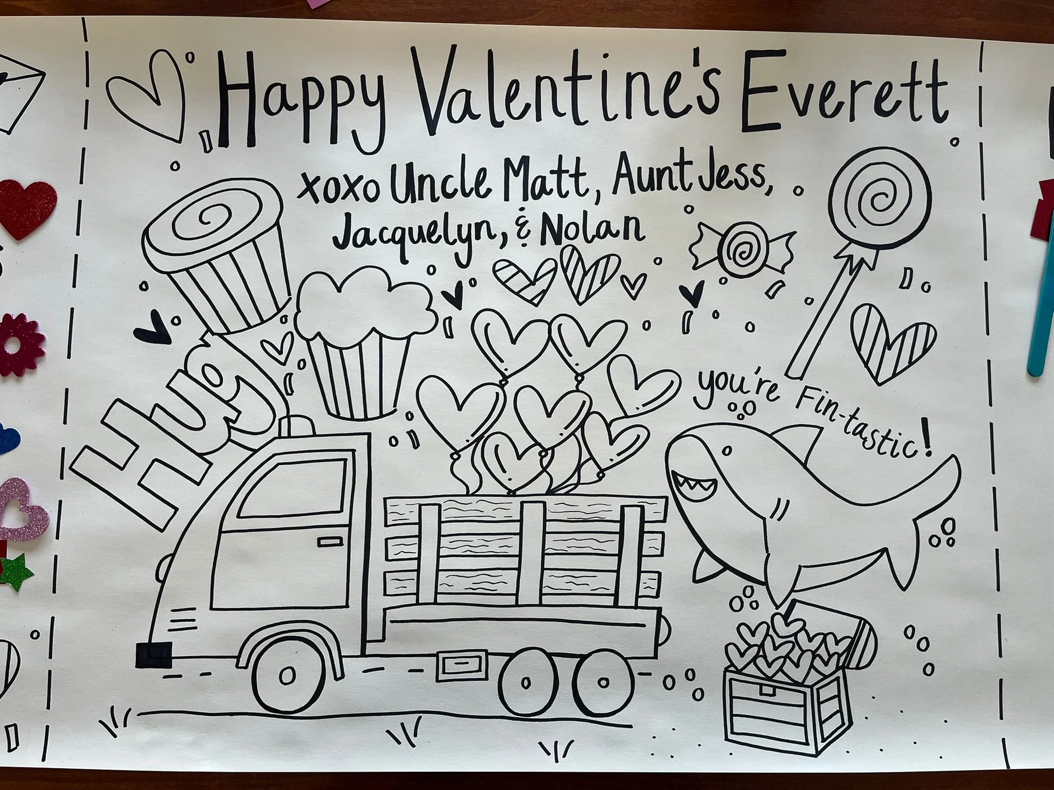 Black and white coloring page with Valentine's Day themed drawings, including a truck carrying presents, balloons, and hearts, along with the greeting 'Happy Valentine's Everett' and a message from family members.