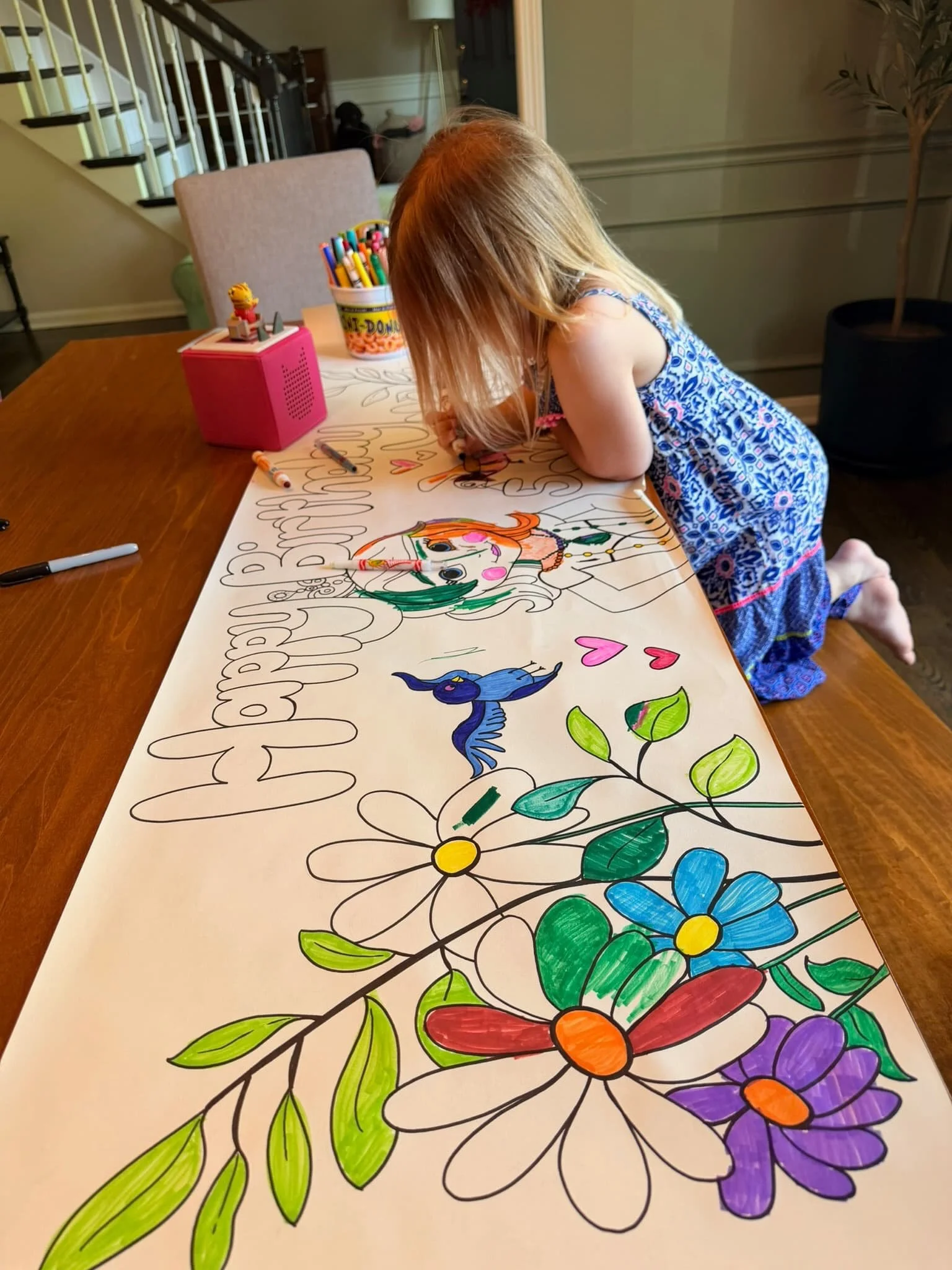 A young girl is coloring a large poster with floral designs and a unicorn. She is using markers to color the flowers and unicorn, and the poster is on a wooden table. There are more markers, a pink speaker, and a container of additional markers on the table.