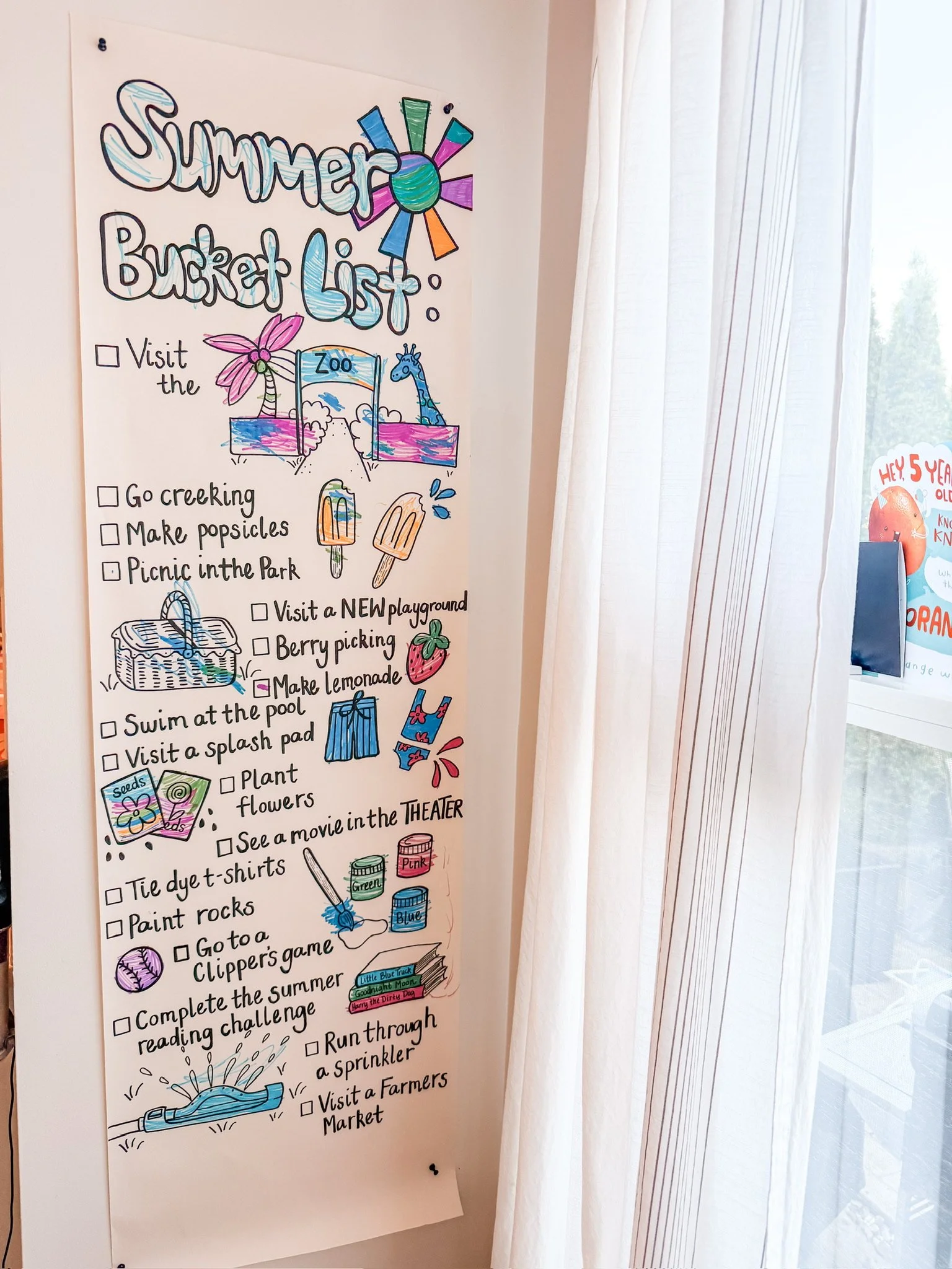 A colorful summer bucket list written on a large white paper with illustrations next to each activity. It includes activities such as visiting the zoo, going creekin, making popsicles, having a picnic, visiting a new playground, berry picking, making lemonade, swimming, visiting a splash pad, planting flowers, and more.
