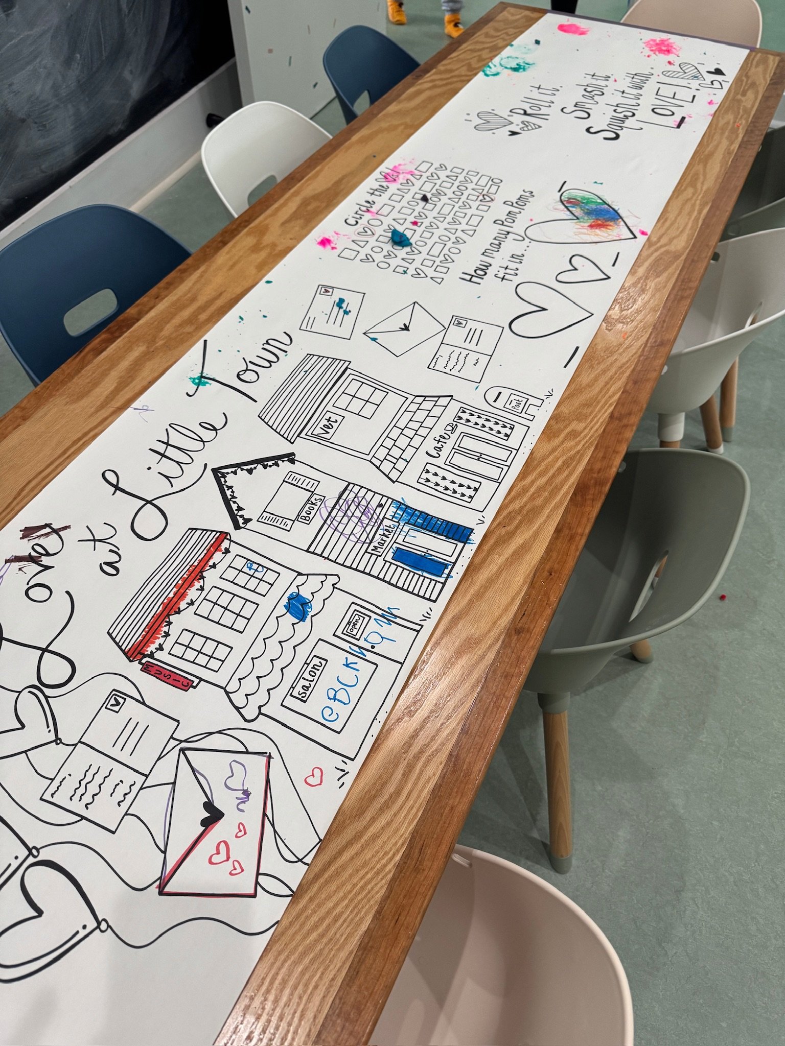 A long whiteboard on a wooden table with colorful drawings and handwritten notes about a community or school project, including a house, mail, and hearts, surrounded by empty chairs.
