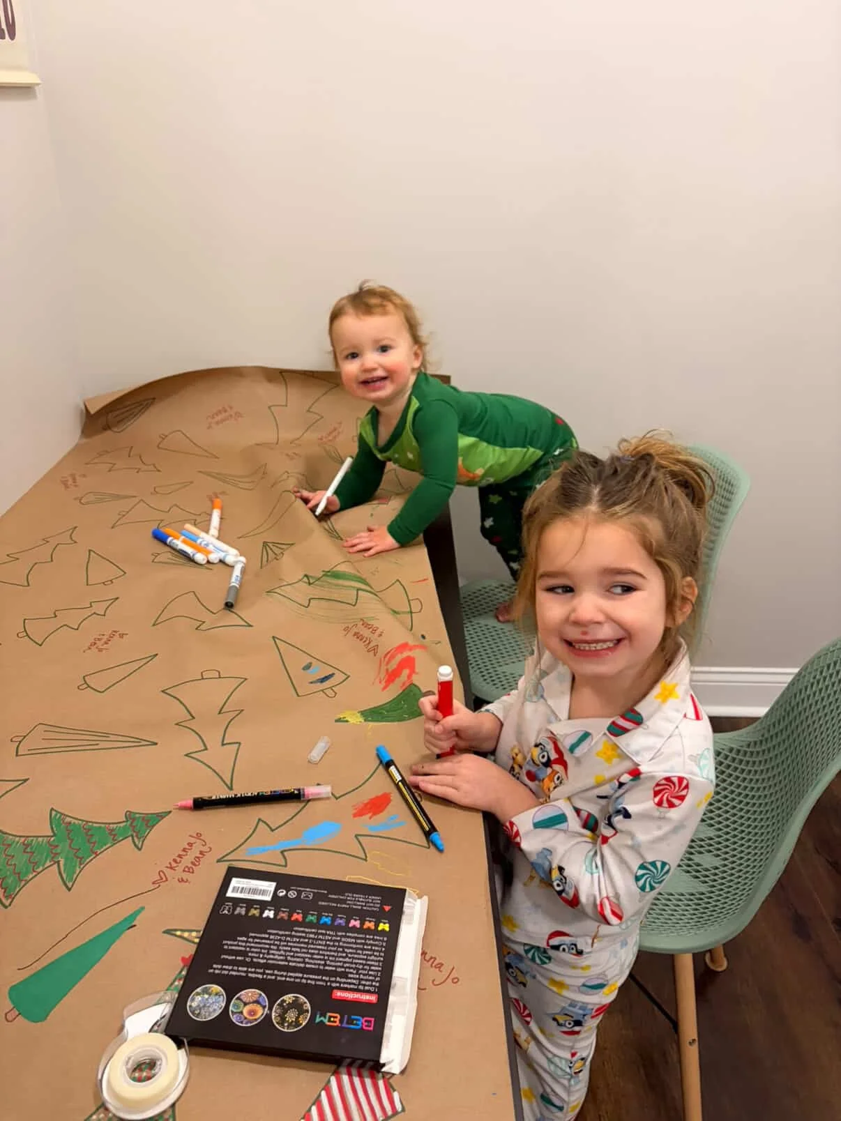 Two young children drawing and coloring Christmas-themed pictures on large paper attached to a table.