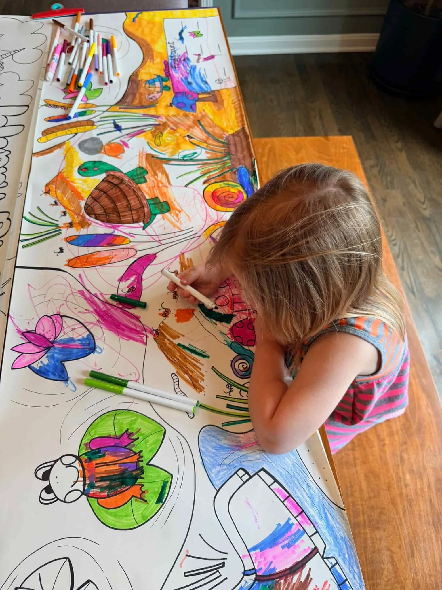 A child coloring a large, detailed poster with various illustrations of nature, animals, and a rainbow using colorful markers. The child is focused on the drawing, which includes a turtle, a butterfly, and other vibrant images.