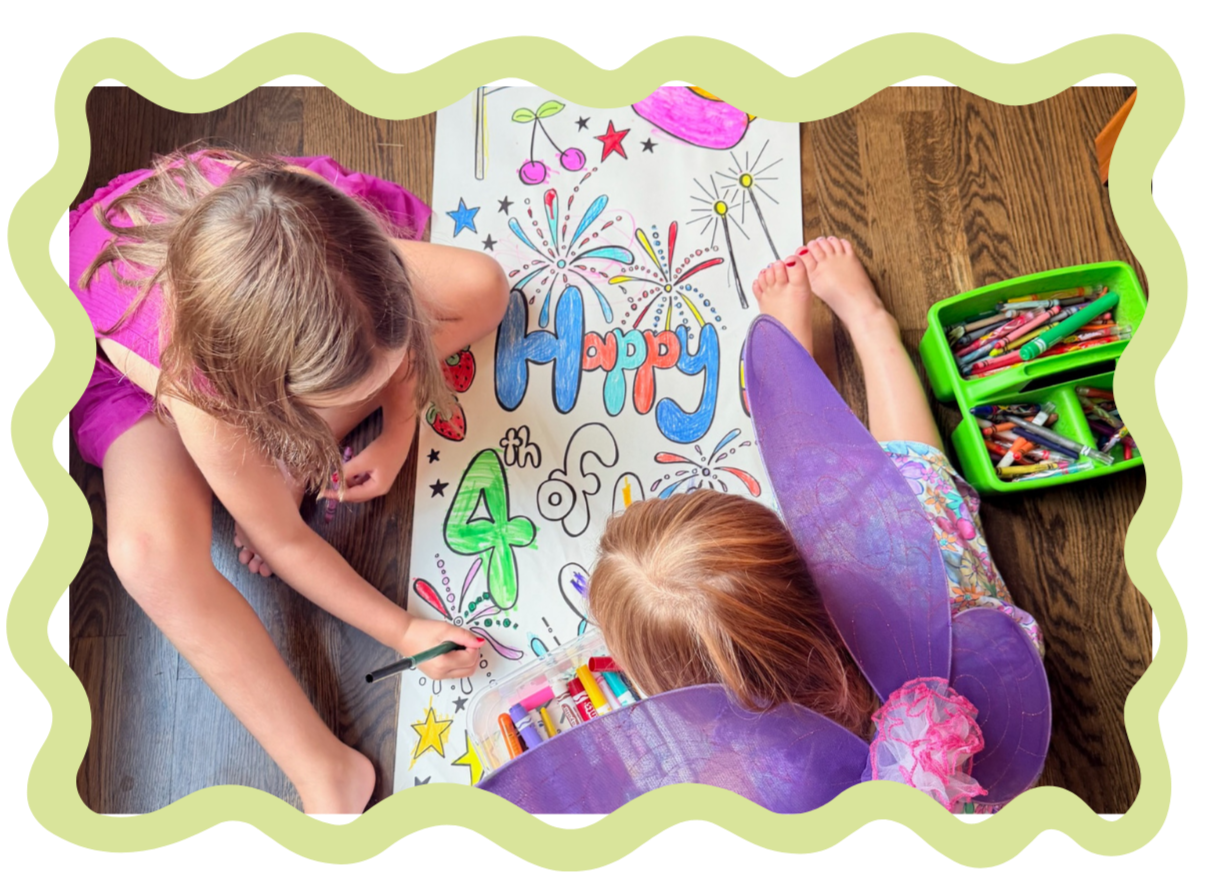 Two children, one with fairy wings, are coloring and decorating a large poster with the words "Happy 4th" surrounded by fireworks, stars, and other festive drawings on a wooden floor.