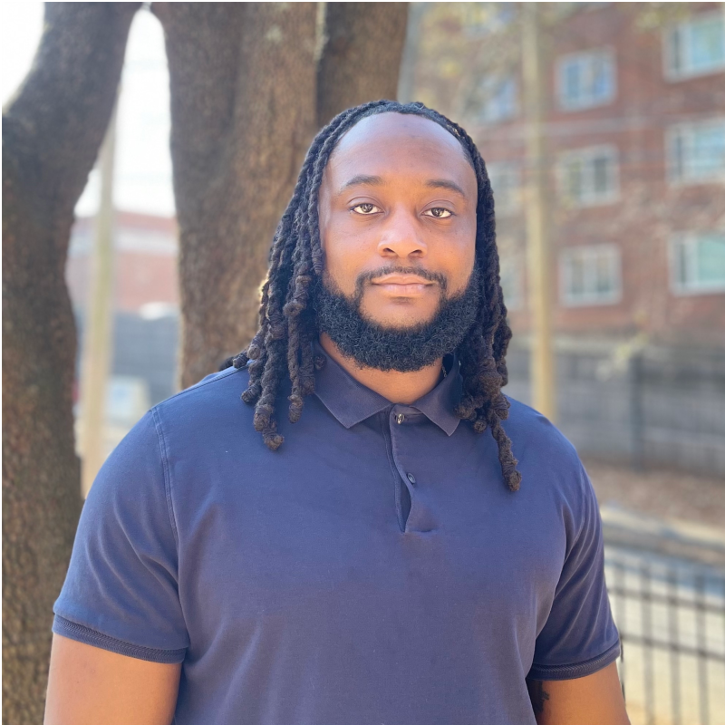 A man with long dreadlocks and a beard standing outdoors in front of trees and a building, wearing a navy blue polo shirt.