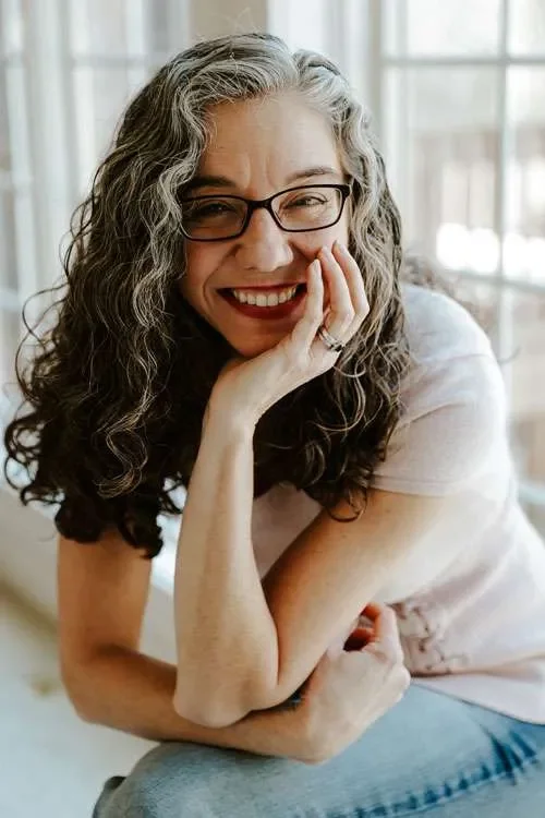 A woman with curly black and gray hair, glasses, and a white shirt smiling while sitting near a window.
