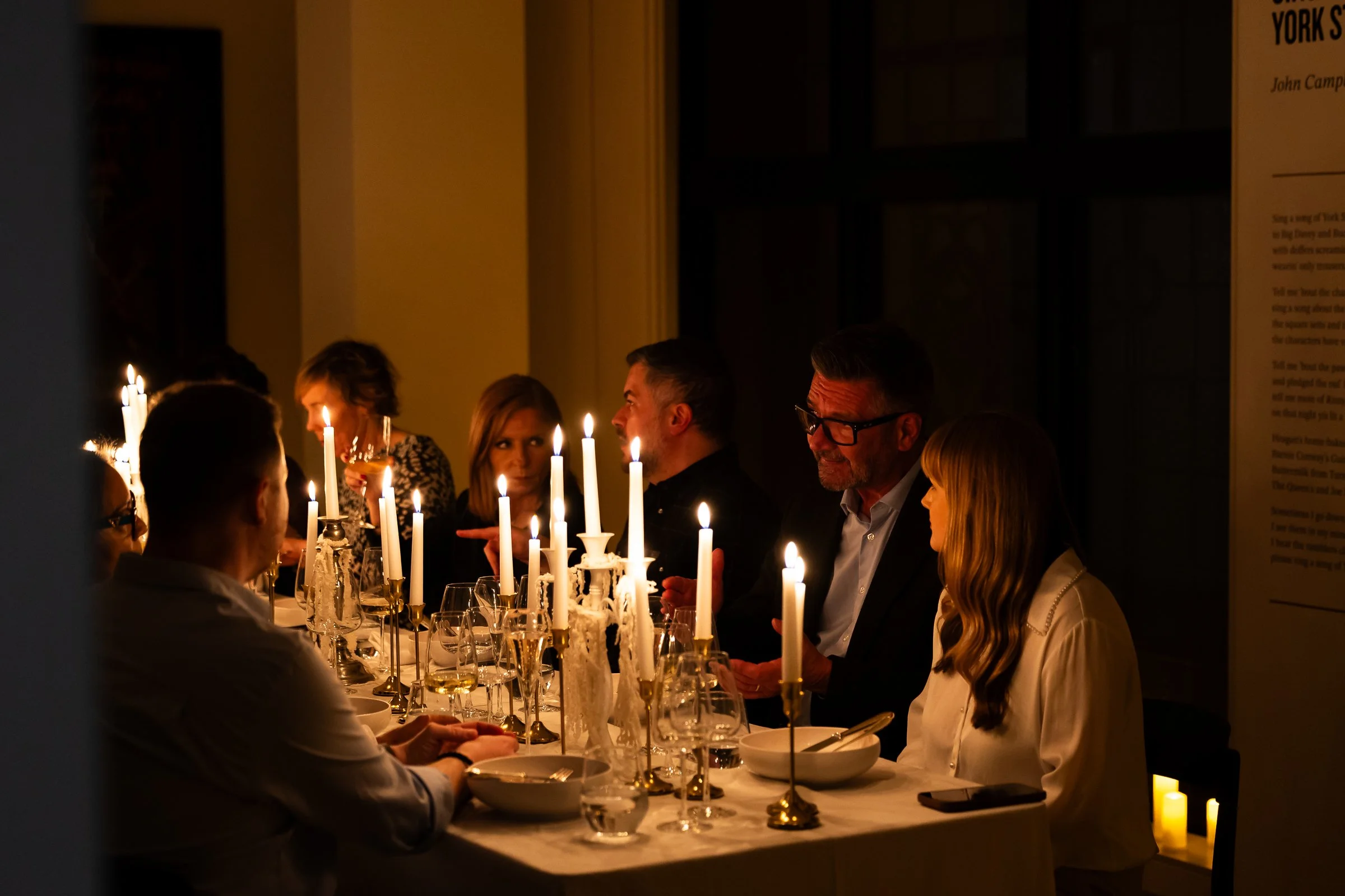 People gathered around a dinner table with lit candles, engaging in conversation, in a dimly lit room.