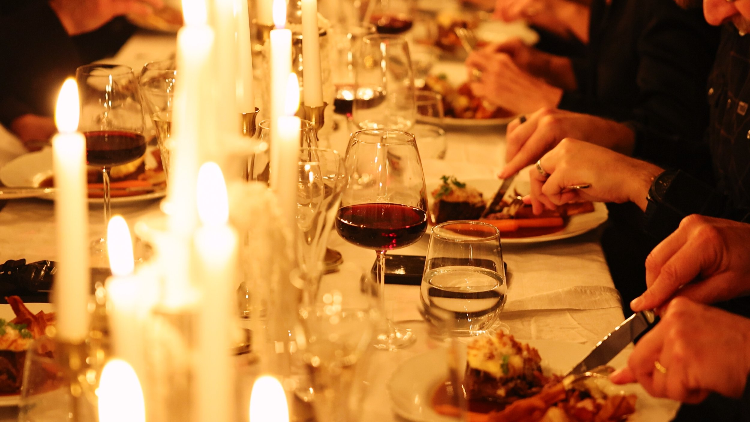 A dinner table with lit candles, wine glasses filled with red wine, and people serving themselves food in a warm, intimate setting.