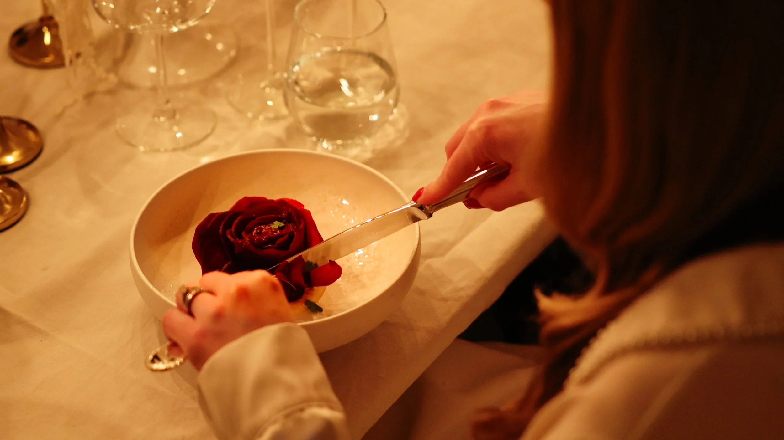 Person cutting a red rose with a knife over a white bowl, with other glasses on the table, in a warm lit setting.