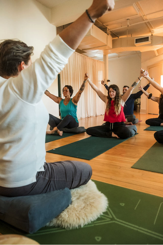 A group of people participating in a yoga or meditation class, sitting on mats with their arms raised, in a well-lit studio with wooden floors and beige curtains.