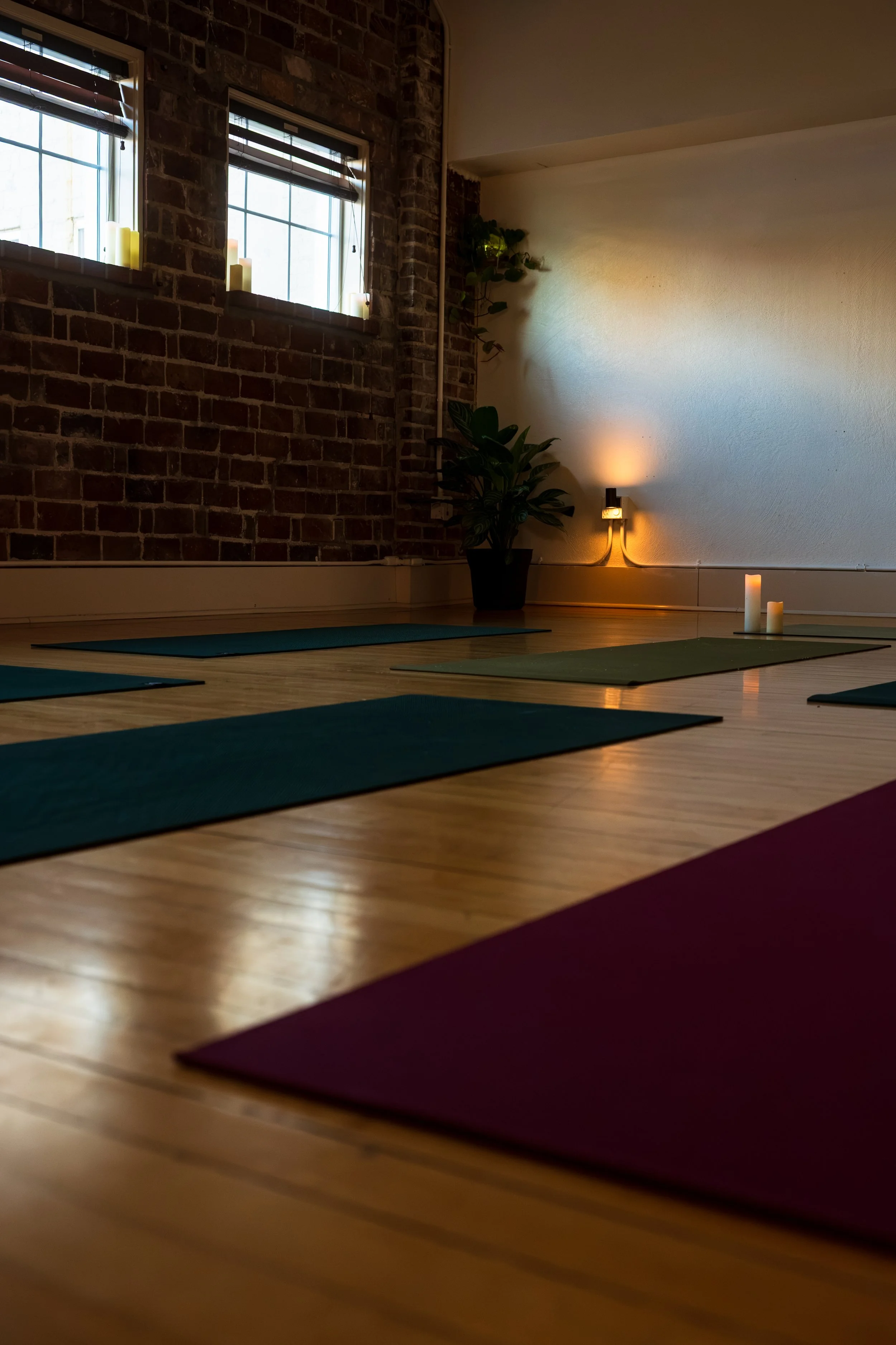 Yoga mats placed on a wooden floor in a dimly lit studio with brick walls, candles, a potted plant, and a wall-mounted light