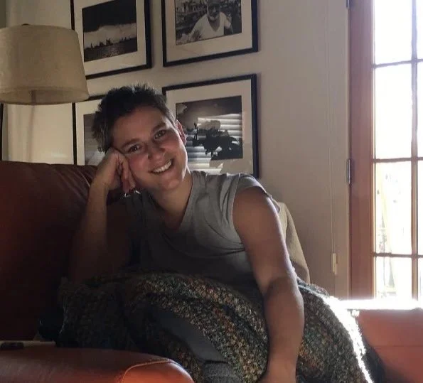 A woman with short hair smiling, sitting on a couch in a living room, with a blanket over her lap, and framed pictures on the wall behind her. Natural light coming in through a window on the right.