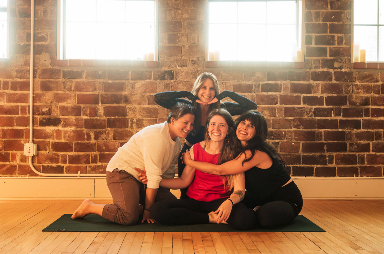 Four women practicing yoga together in a studio with brick walls and large windows, sitting on a yoga mat and smiling.