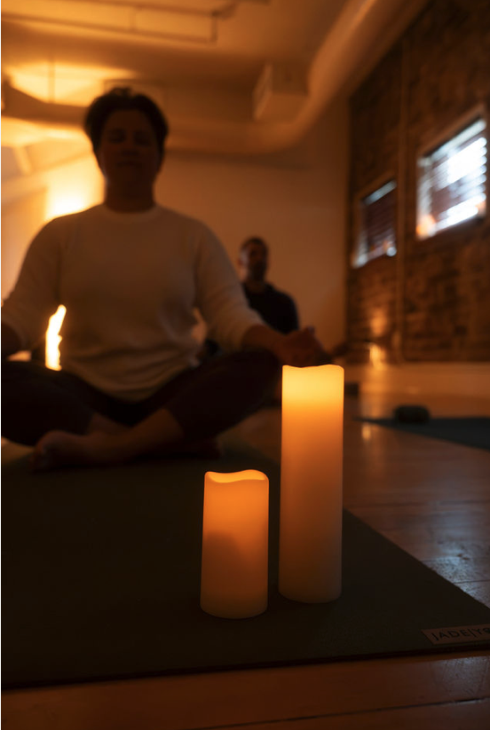 Two people practicing yoga or meditation indoors with candles and a yoga mat in a cozy, softly lit room.