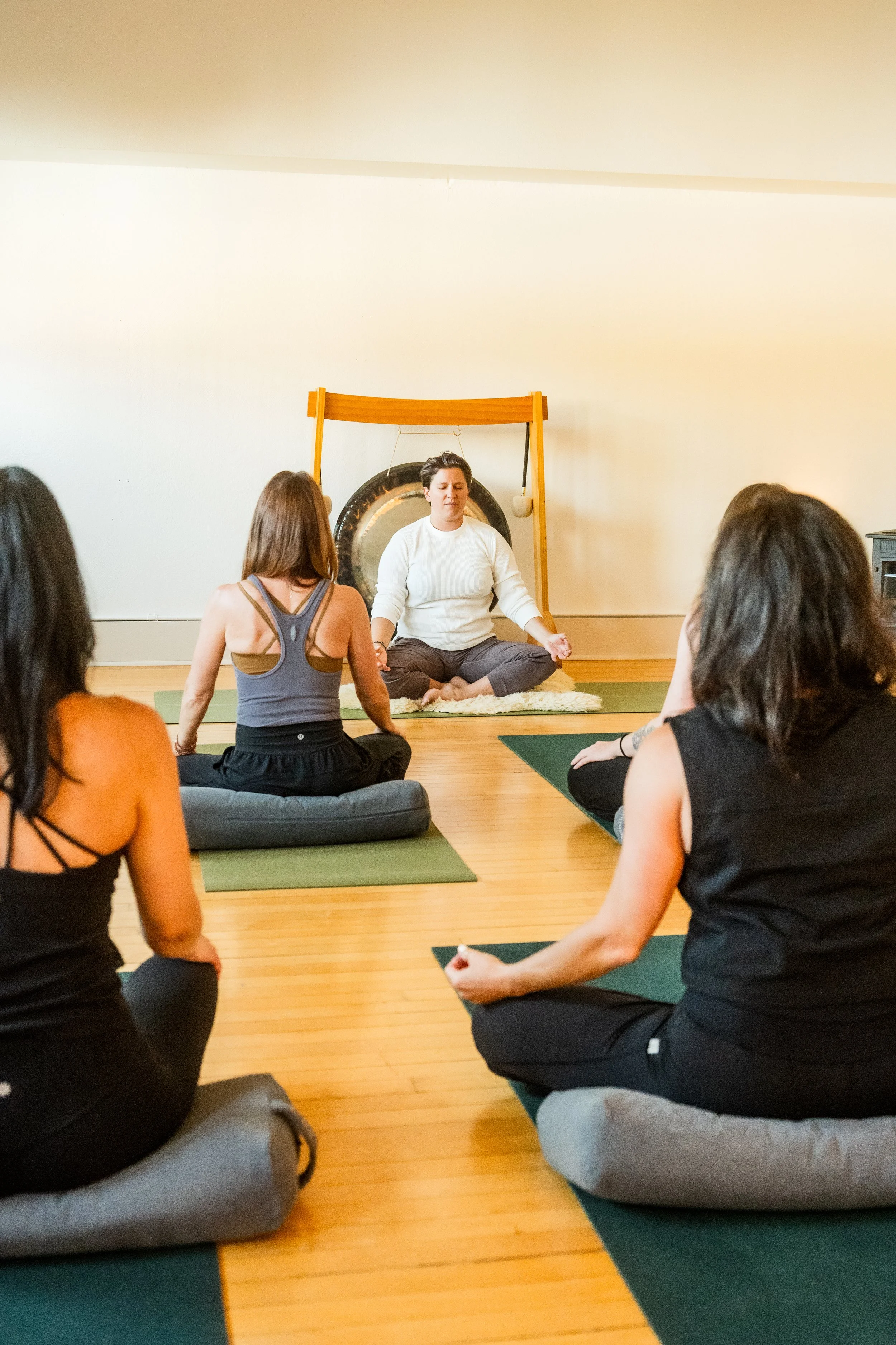 People participating in a yoga class, seated on mats in a meditation pose, with a woman instructor sitting cross-legged at the front, in a room with wooden flooring and plain walls.