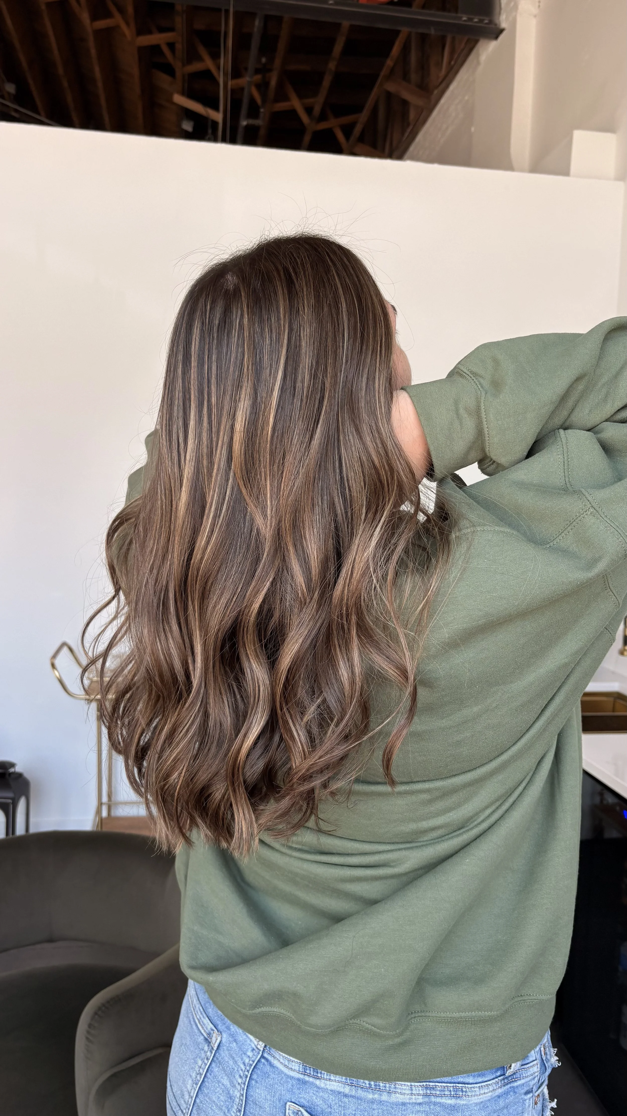 A woman with long, wavy brown and blonde hair wearing a green sweatshirt and blue jeans, standing in a room with a white wall, a modern brown chair, and visible ceiling beams.