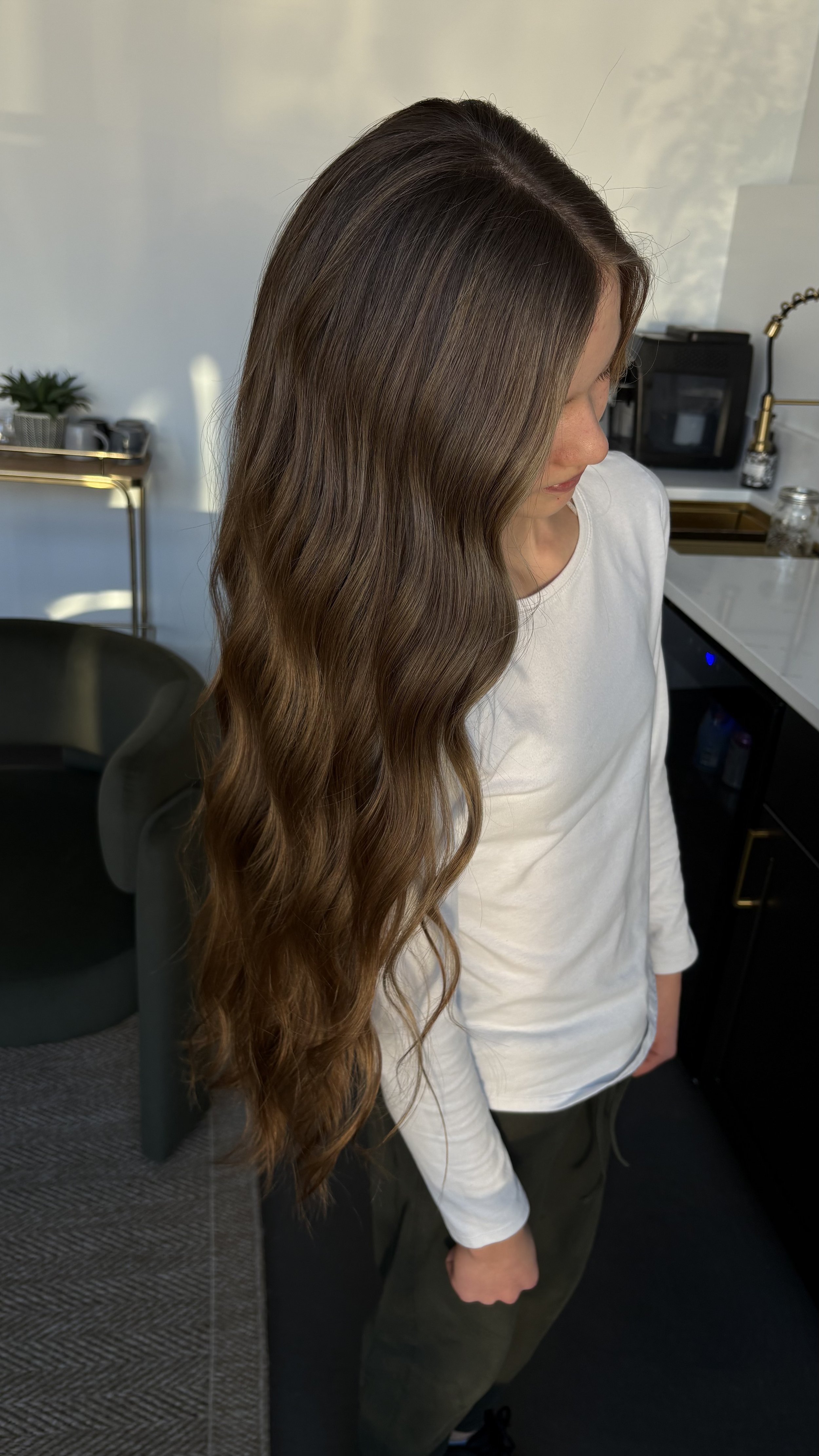 A woman with long, wavy brown hair wearing a white shirt and green pants standing in a room with kitchen appliances and furniture.