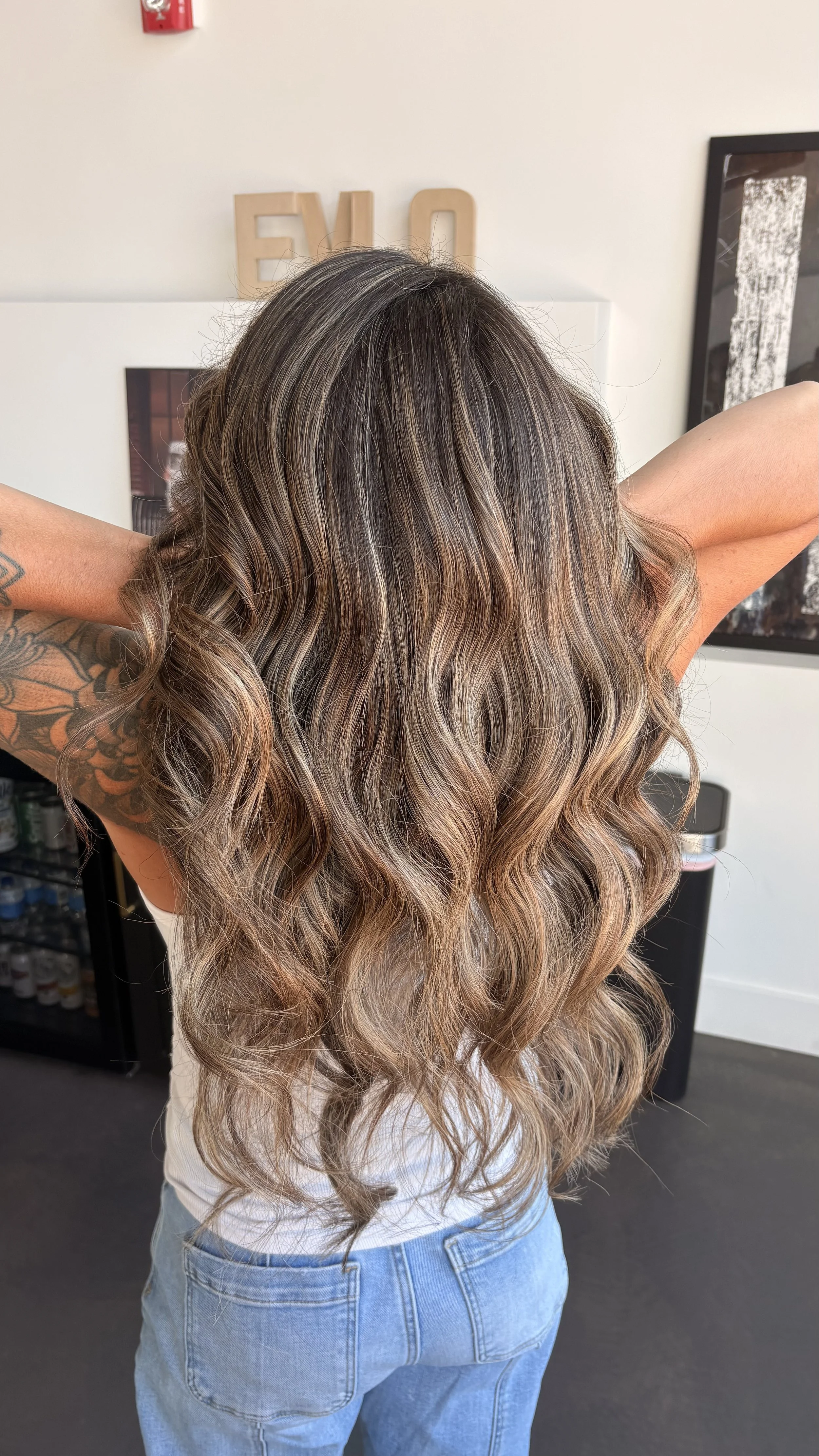Person with long, wavy, highlight-colored hair wearing blue jeans, standing in a salon with arms raised.