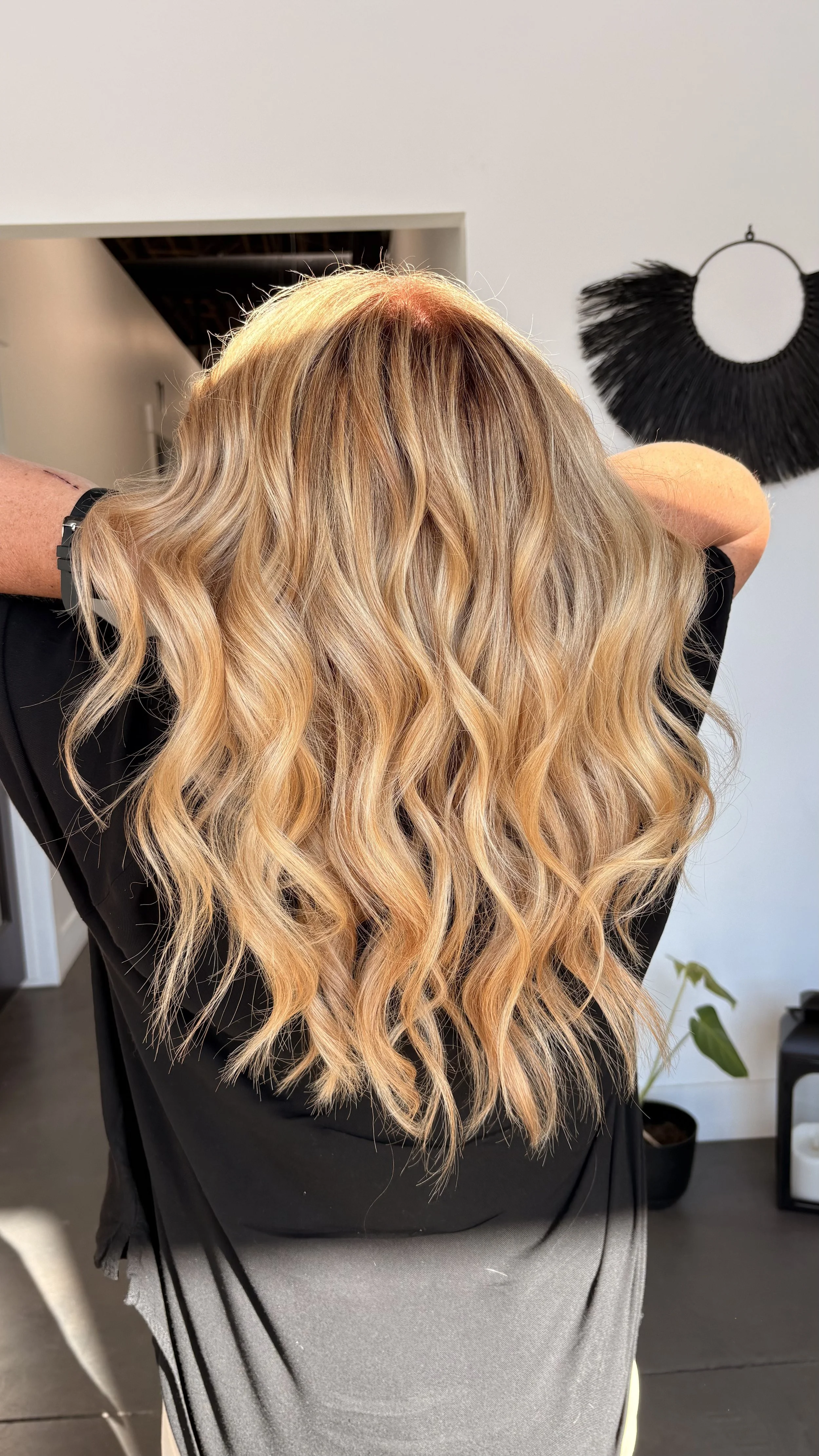 A person with long, wavy, blonde hair wearing a black top and gray skirt, with their back facing the camera. There is a black decorative wall hanging in the background.