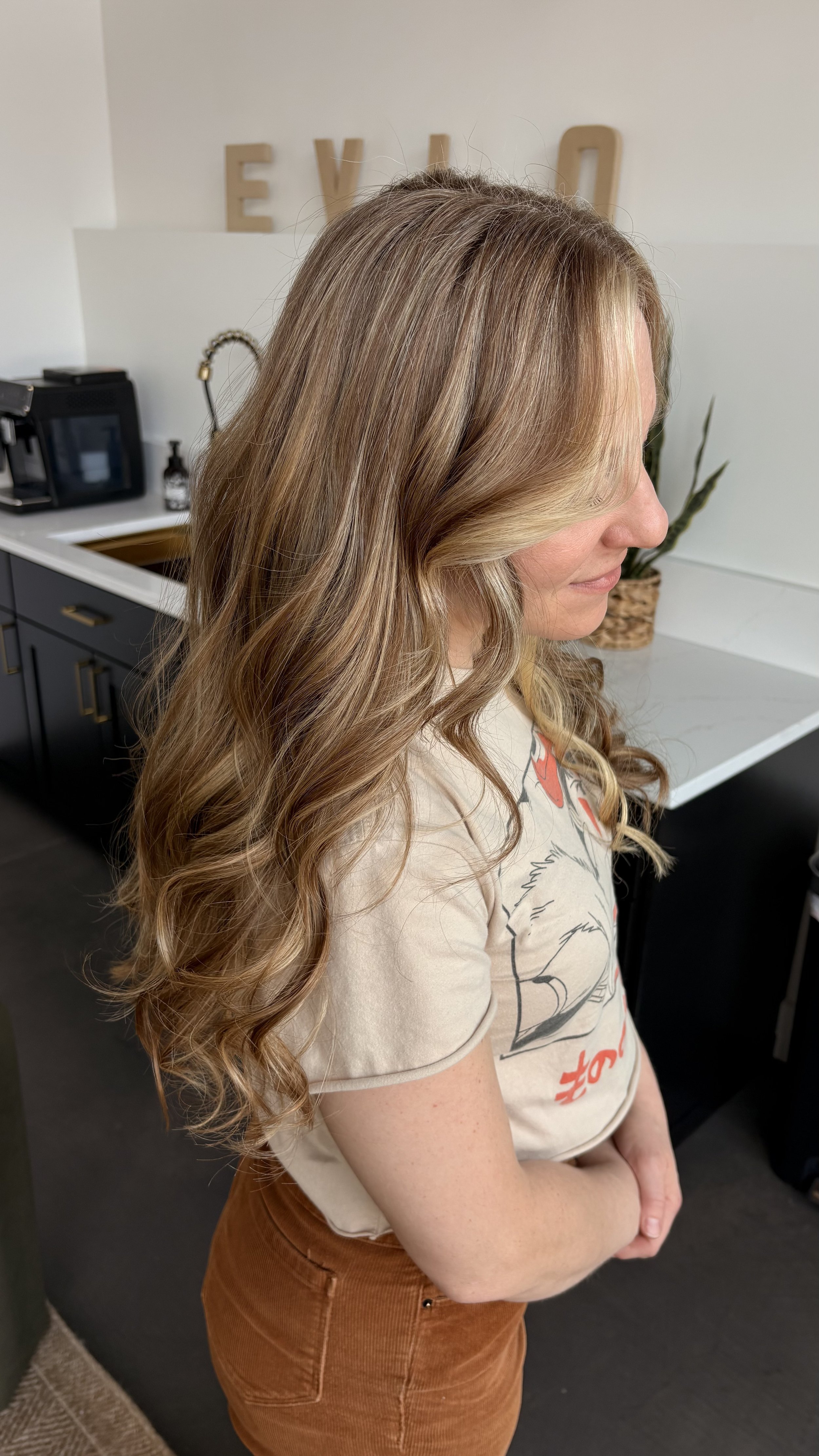 A woman with long, wavy blonde hair standing sideways in a kitchen, wearing a beige T-shirt and rust-colored pants.