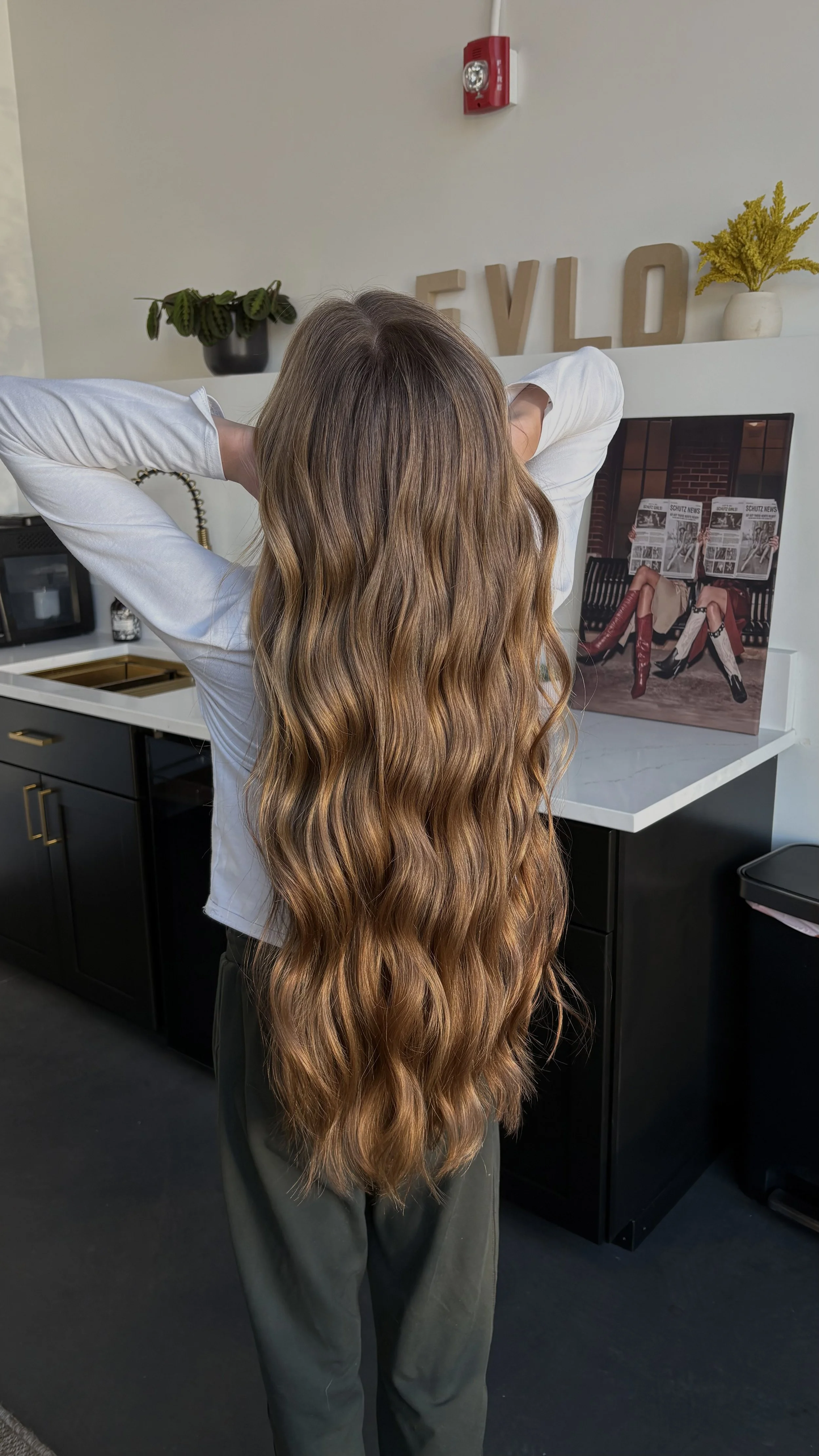 A person with long wavy brown hair standing in an indoor setting, facing away from the camera. They are wearing a white top and dark pants. Behind them, there is a counter with a plant, decorative letters spelling 'EVOLO', and a framed photo of two p