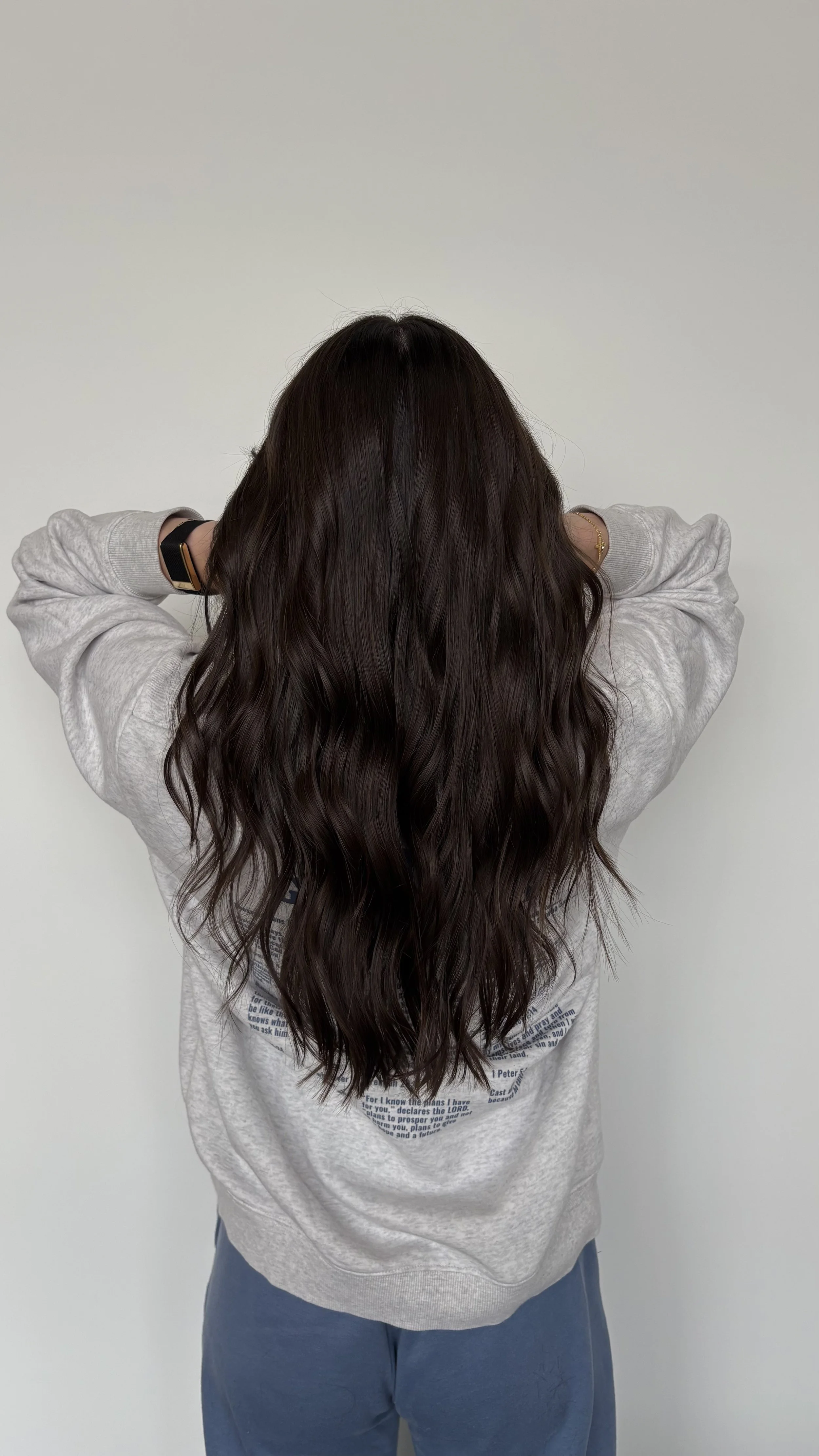 Back view of a woman with long, wavy, dark brown hair, wearing a light gray hoodie and blue sweatpants, standing against a plain white wall.