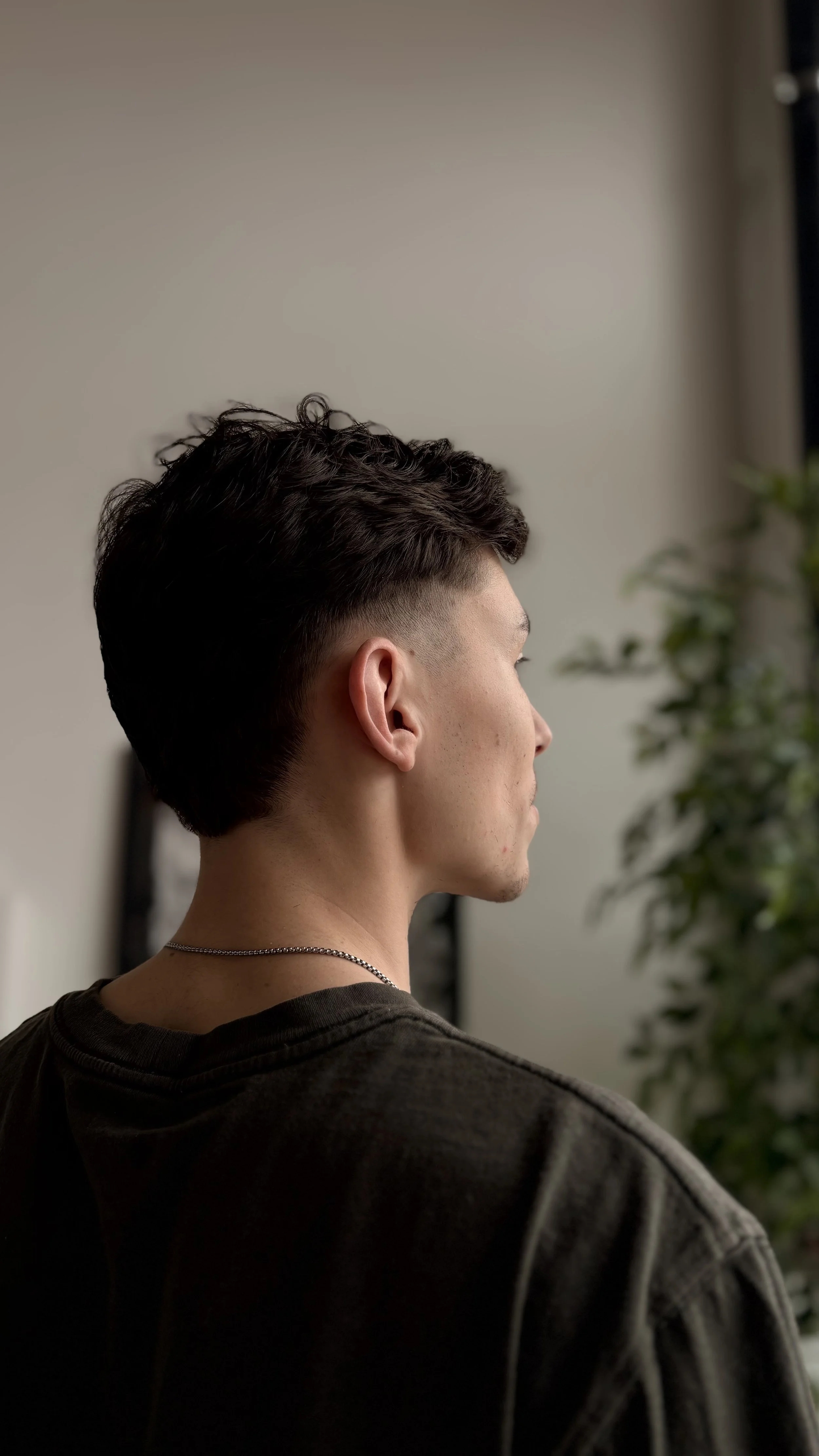 Side profile of a young man with short, dark, wavy hair, wearing a dark shirt and a thin silver necklace, standing indoors with a blurred plant in the background.
