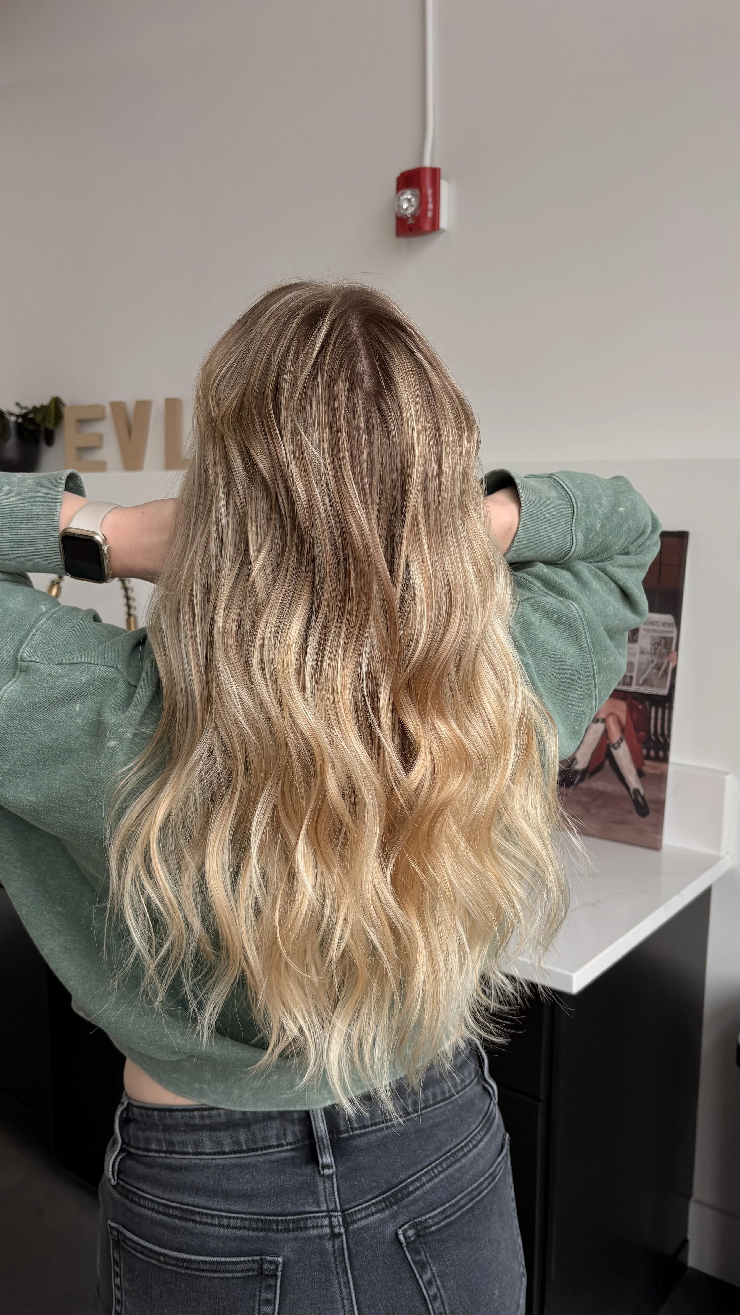 A person with long, wavy blond hair wearing a green sweatshirt and dark jeans, standing with their back to the camera in an indoor setting.