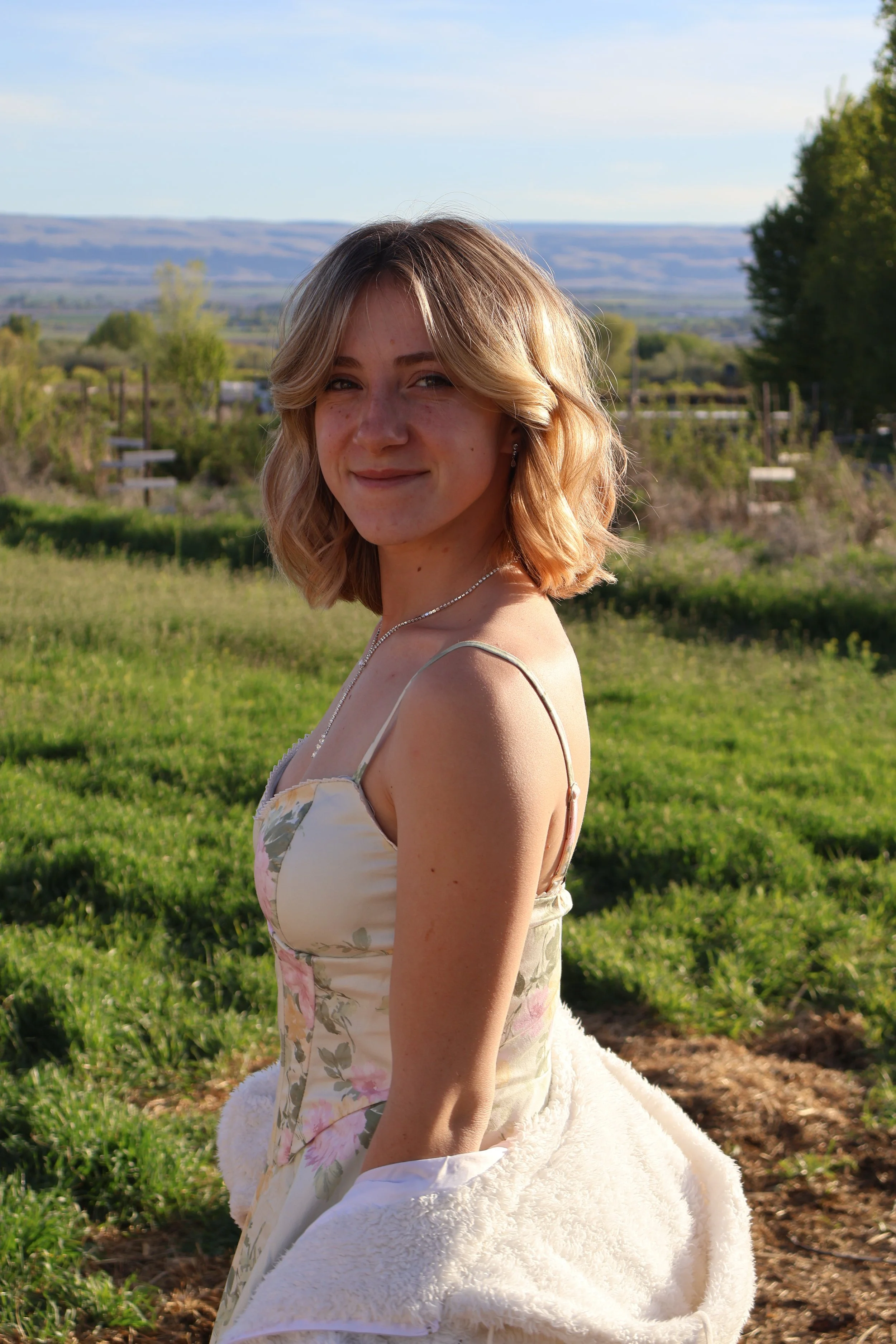A young woman with shoulder-length blonde hair wearing a floral dress and jewelry, smiling outdoors in a scenic landscape with green fields, trees, and distant hills under a clear sky.