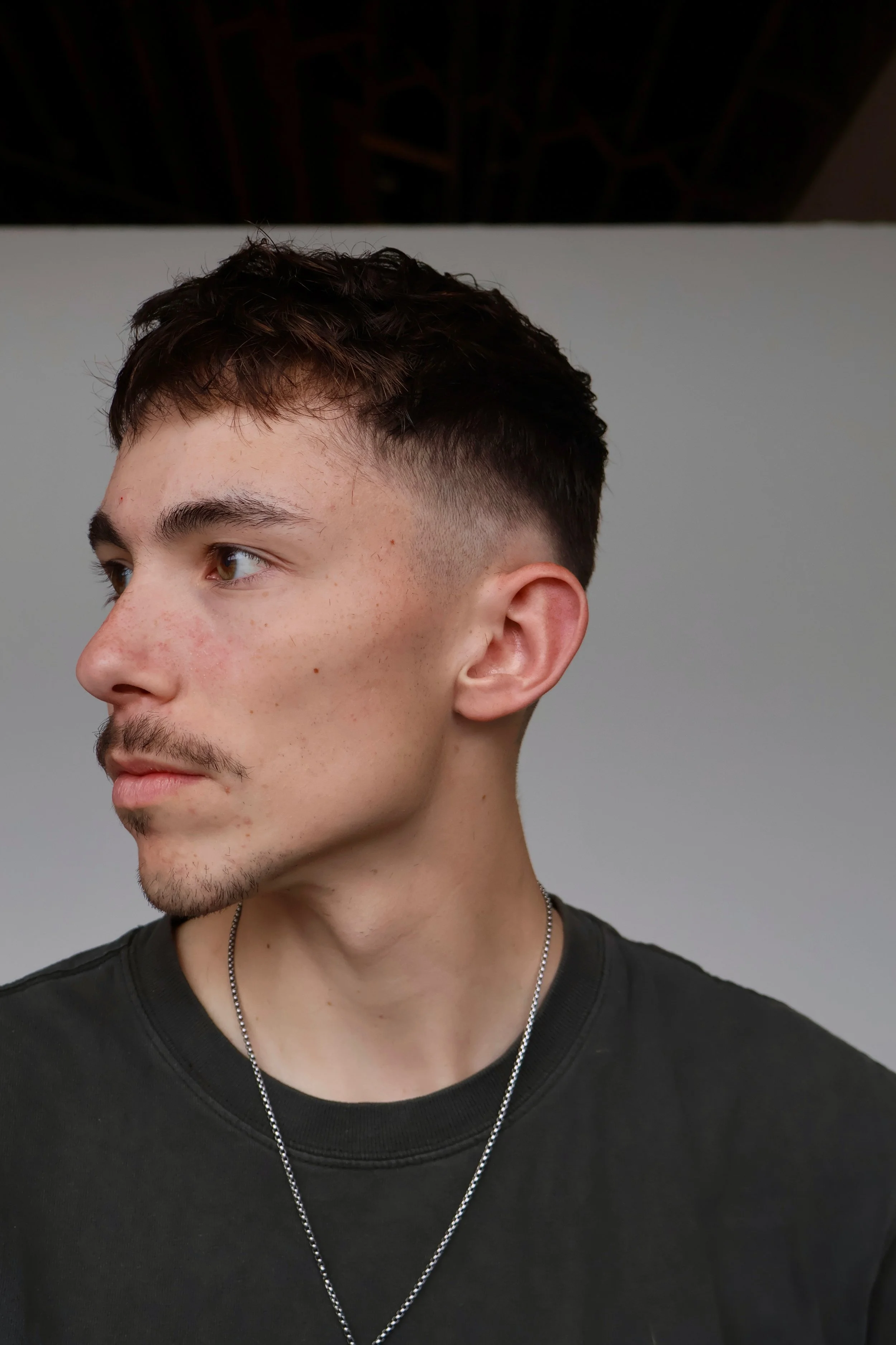 A young man with short dark hair, brown eyes, light skin, and facial hair, wearing a black shirt and a silver chain necklace, is facing left against a plain background.
