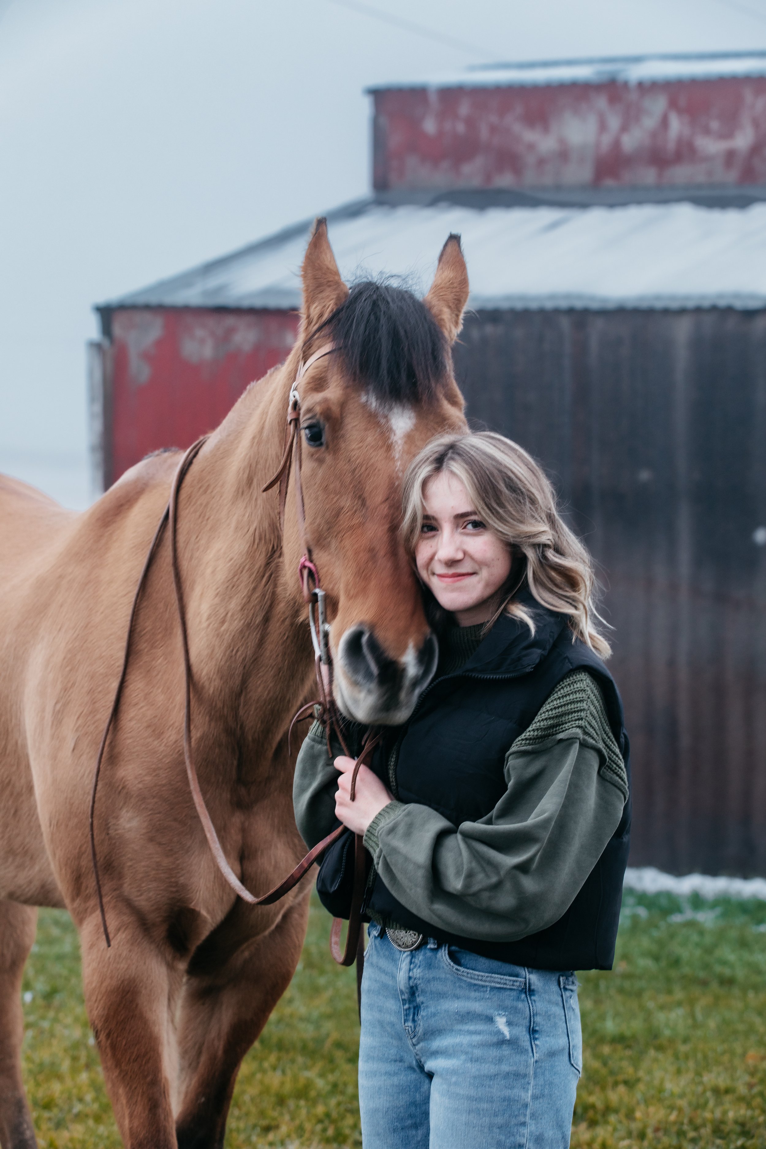 A young woman with blonde curly hair and a black and green jacket standing outside with a brown horse on a cloudy day.