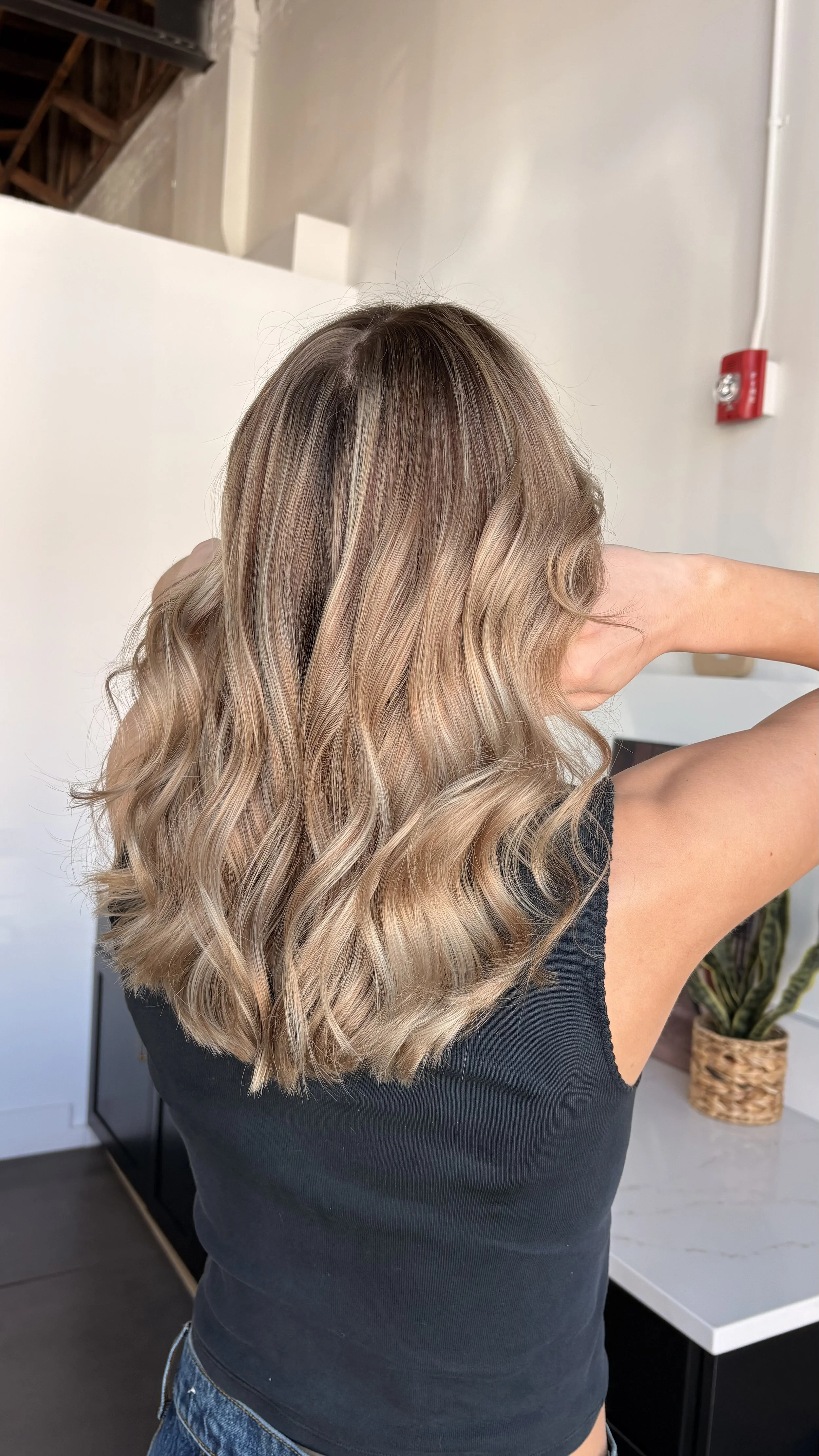 Woman with long, wavy blonde hair in a hair salon, standing with her back to the camera, while wearing a black tank top and blue jeans.