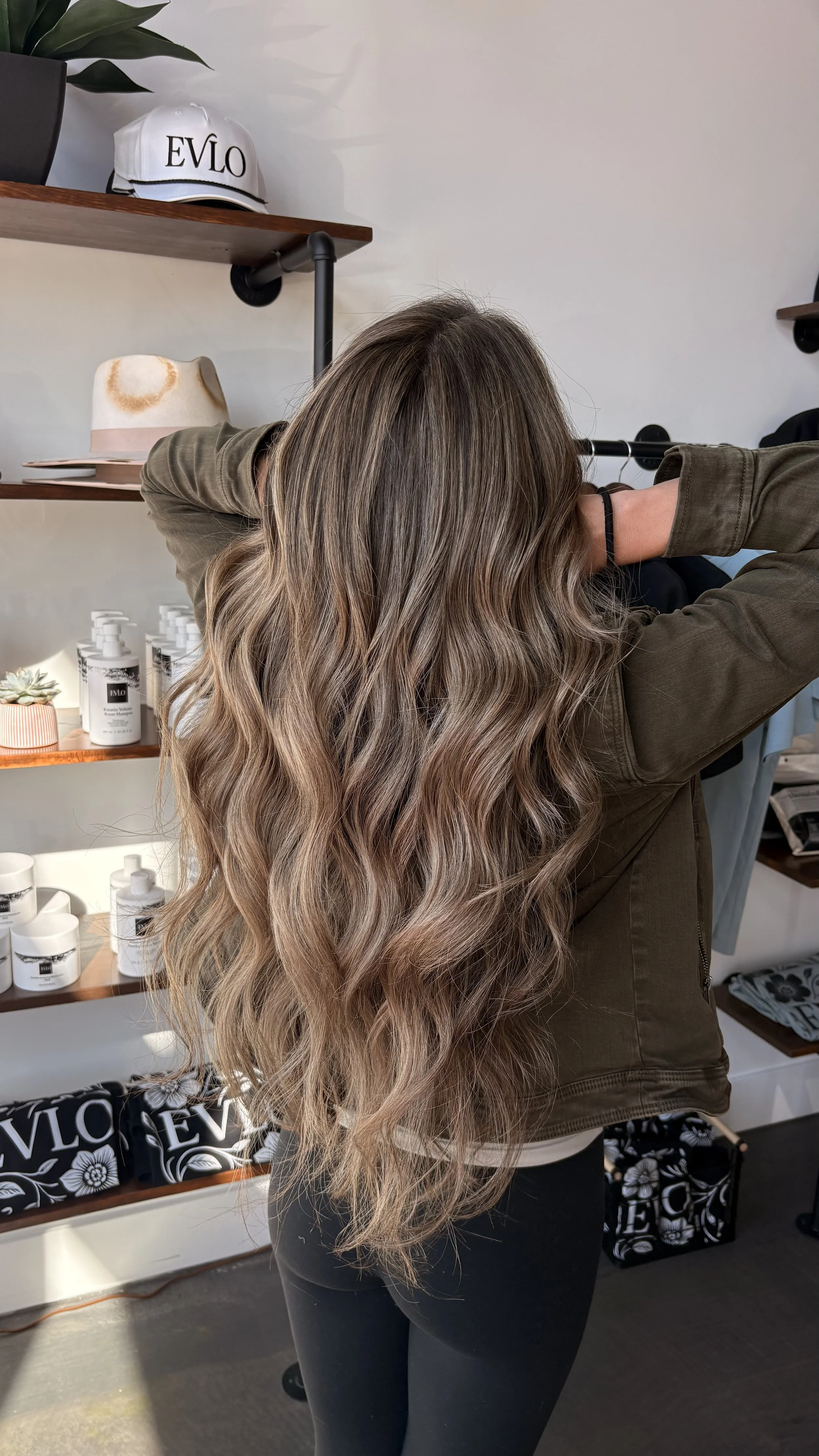 A woman with long, wavy, ash-blonde hair stands with her back to the camera in a salon, with shelves of hair products and a white hat on display in the background.