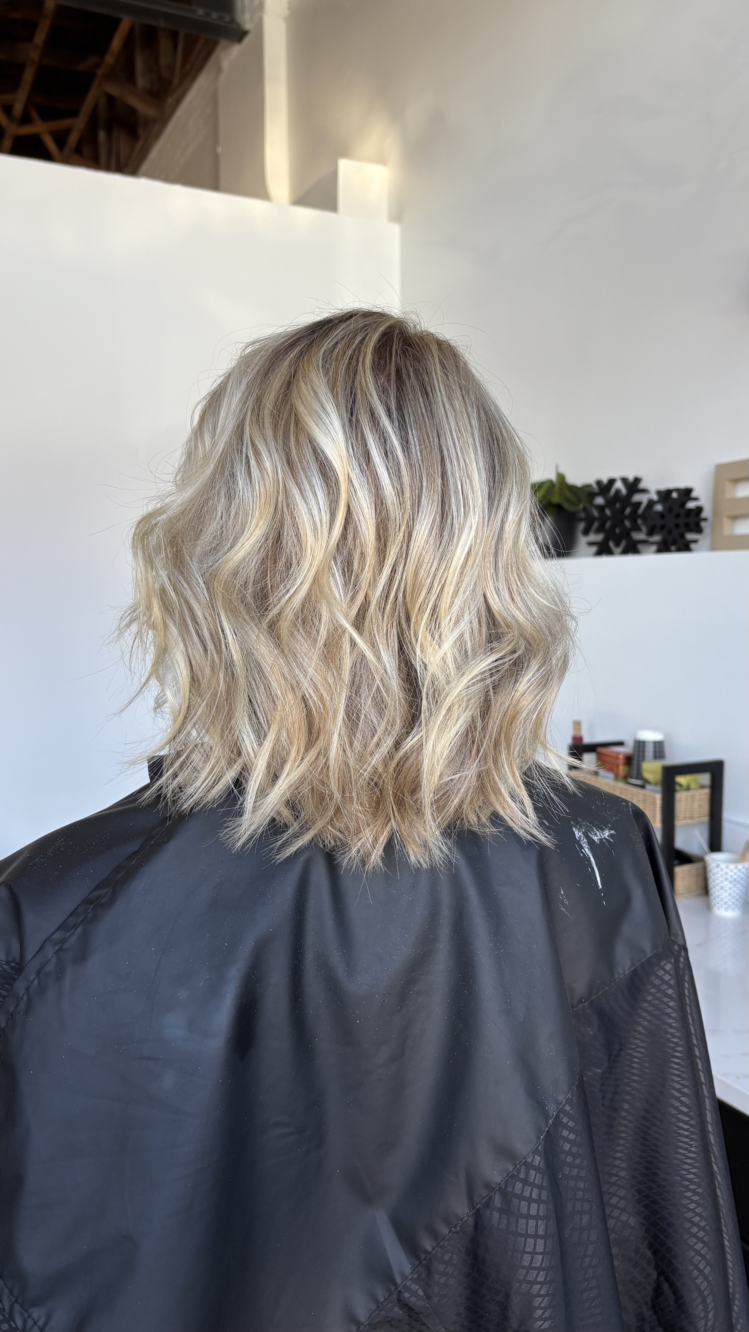 Back of a person with shoulder-length wavy blonde hair, sitting in a salon chair with a black cape, in a salon with white walls and salon supplies in the background.