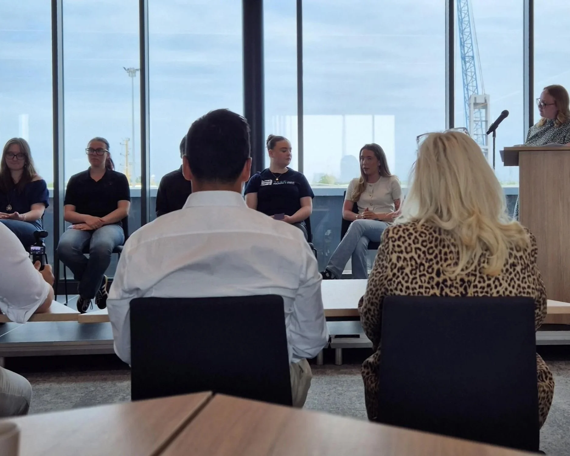 A group of people seated in a modern conference room, facing a woman standing at a podium near large windows with a cityscape view. The attendees include women, some wearing casual or business casual attire, and are listening attentively.