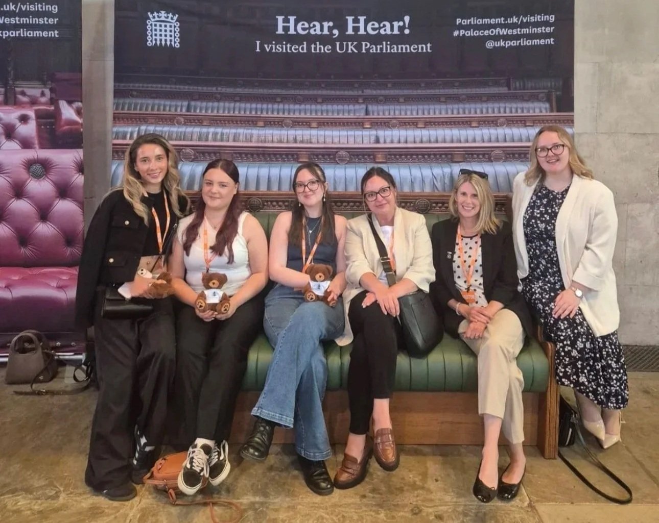 A group of six women posing together at the House of Commons, seated and standing in front of a UK Parliament display. Several members of the group are holding teddy bears and wearing visitor lanyards.