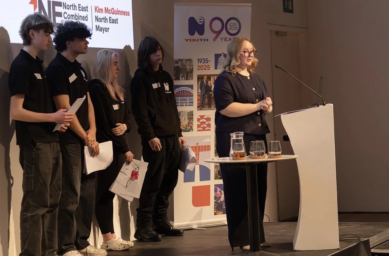 Four young people standing on stage alongside a woman speaking at a podium, with North East Combined Authority and youth event displays in the background.