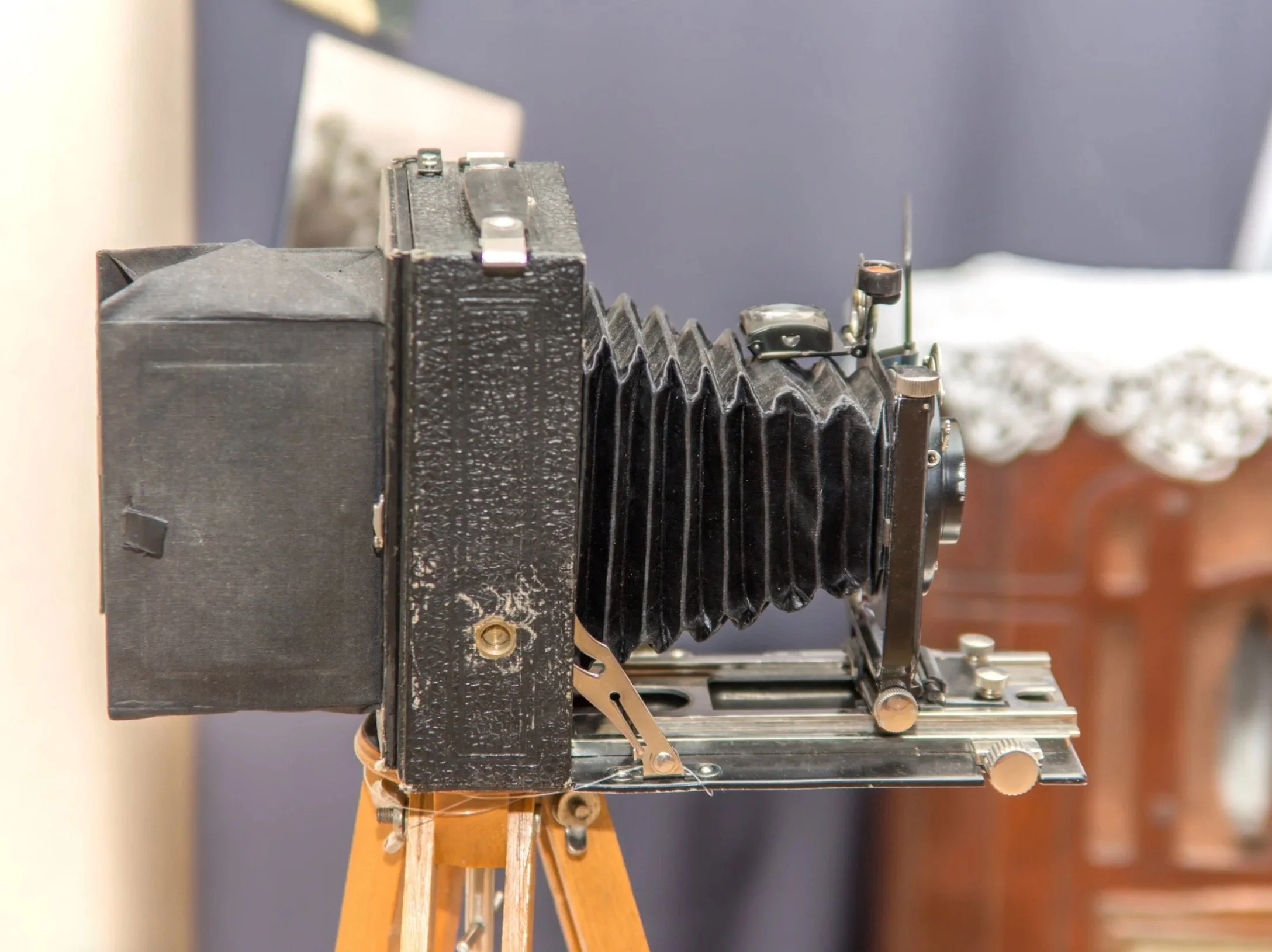 Close-up of an antique large format camera mounted on a wooden tripod, with parts of the camera and tripod visible, against a blurred background.