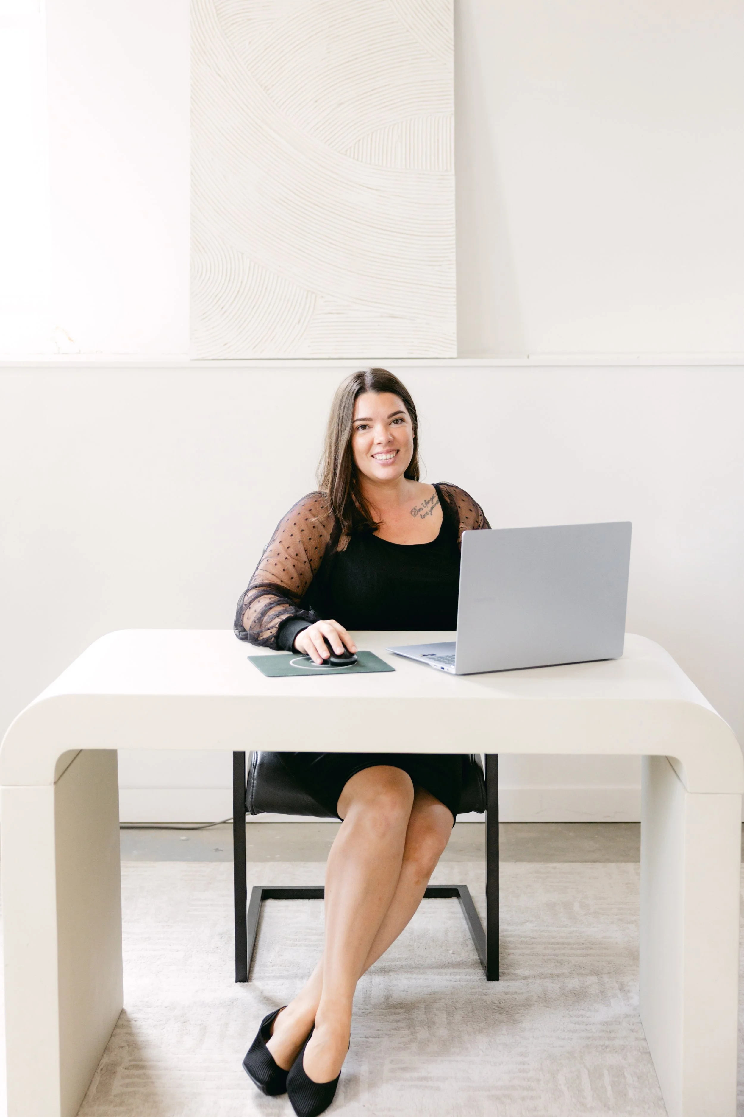 A woman sitting at a white desk in an office, using a laptop and mouse, smiling at the camera. She has dark hair and is wearing a black dress with sheer polka-dot sleeves and black high heels. The room has minimalist decor with a textured abstract art piece on the white wall behind her.