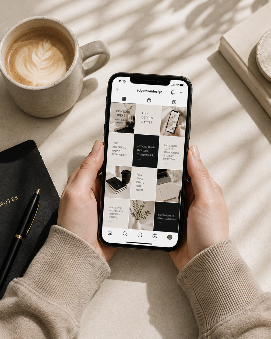 Person holding a smartphone with an Instagram profile open, sitting at a desk with a cup of coffee, a black notebook with a gold pen, and some books.