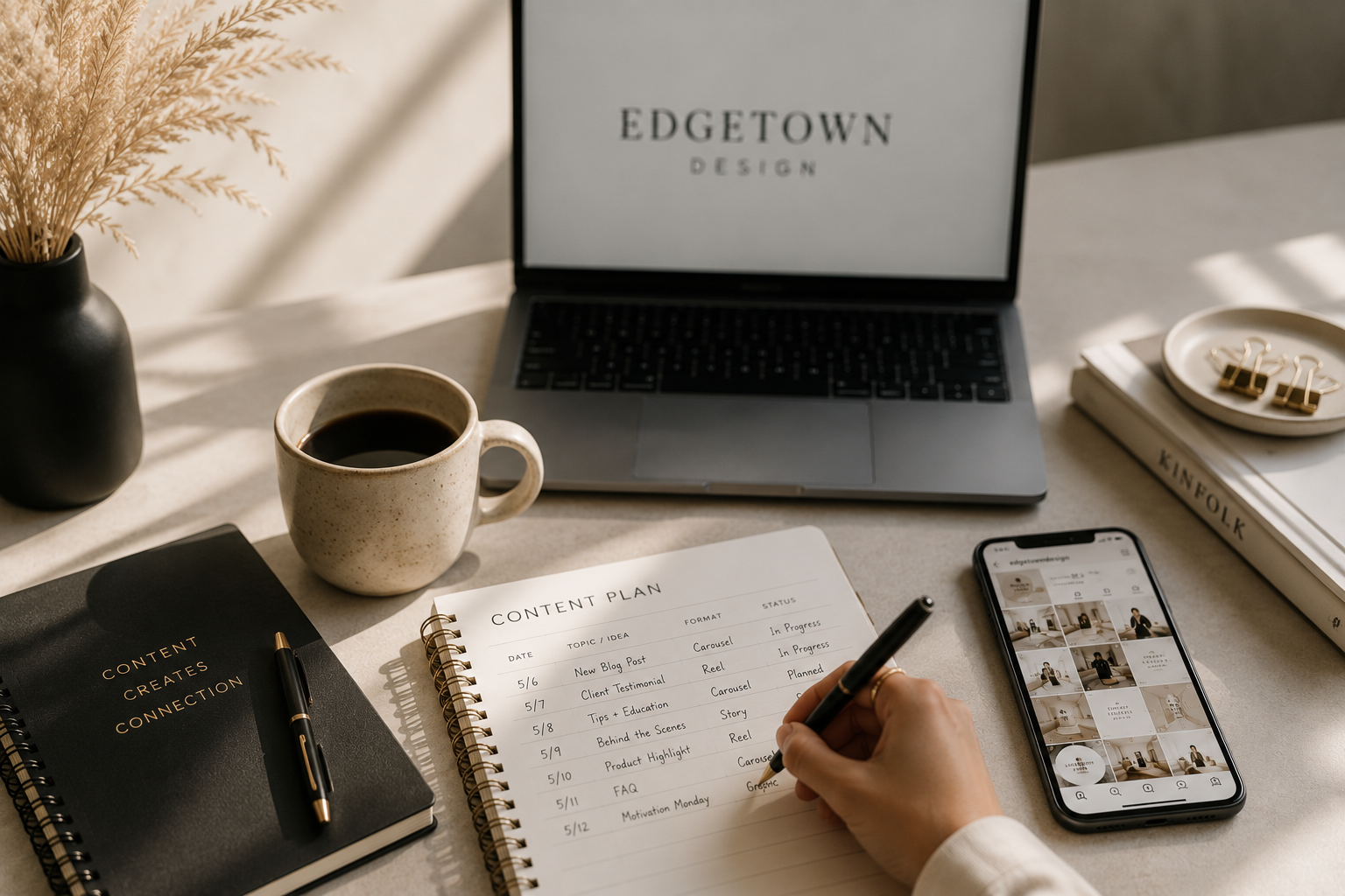 Desk workspace with a coffee mug, a closed black notebook with a pen, a content plan notebook, a laptop displaying 'Edgetown Design,' a smartphone showing a social media grid, a stack of books including a 'Kinfolk' magazine, a small dish with gold binder clips, and a hand writing in the content plan notebook.