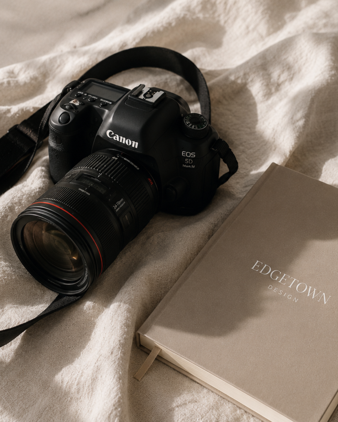 A Canon EOS SD Mark IV digital camera with a black strap, positioned on a cream-colored fabric surface next to a closed beige notebook titled EDGE TOWN DESIGN.
