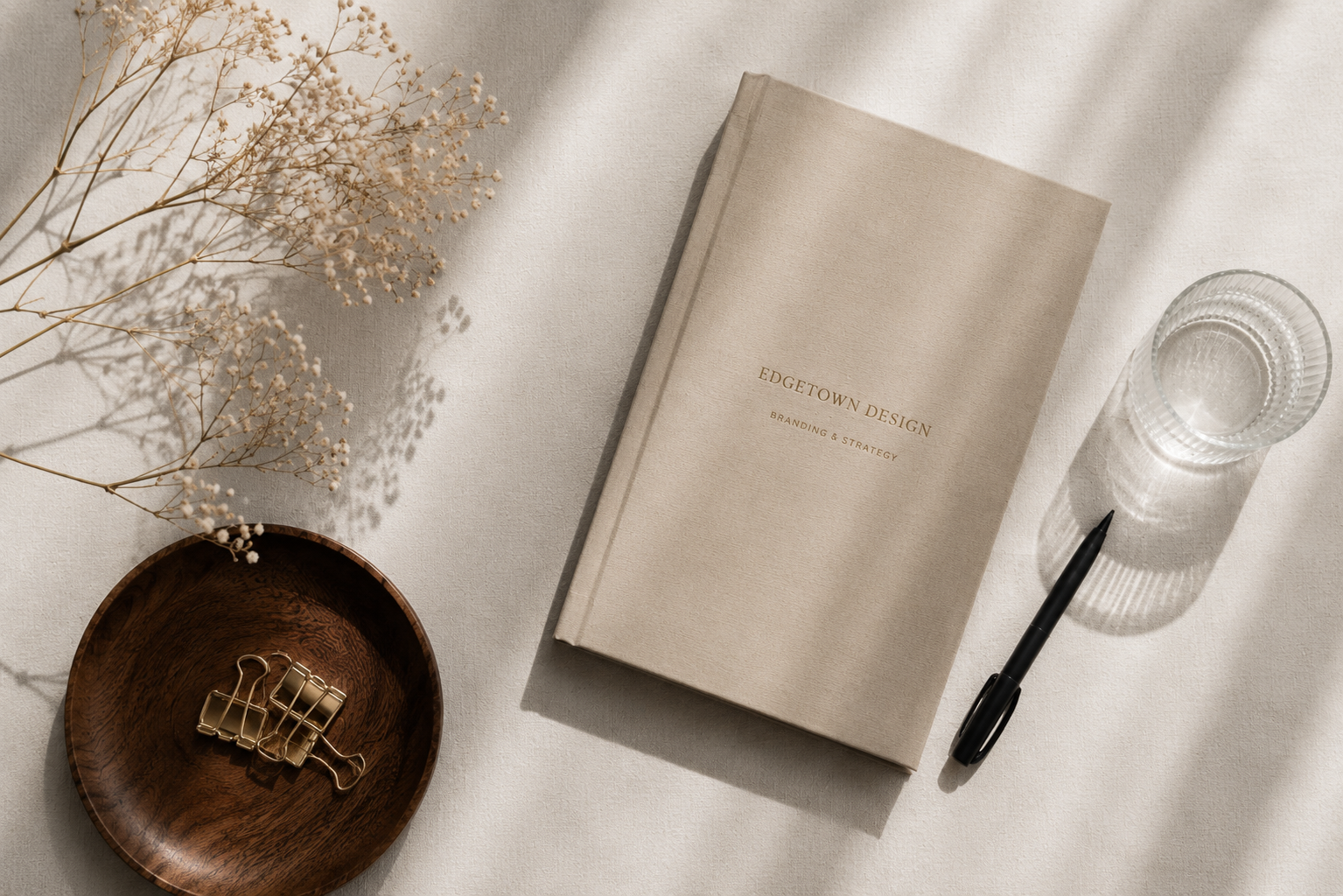 A beige hardcover book titled 'Edgetown Design Branding & Strategy' on a white surface, with a black pen beside it, a glass of water, dried flowers, and a small wooden bowl with gold binder clips. Shadows cast across the surface.