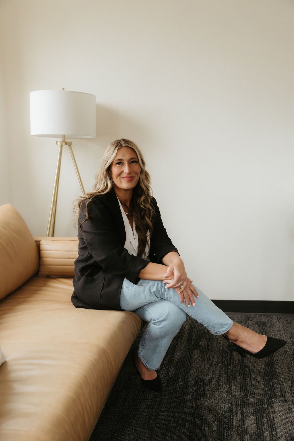 A woman with long wavy blonde hair sitting on a tan leather couch, smiling, wearing a black blazer, white blouse, light blue jeans, and black heels, in a room with a white wall and a tall white lamp.