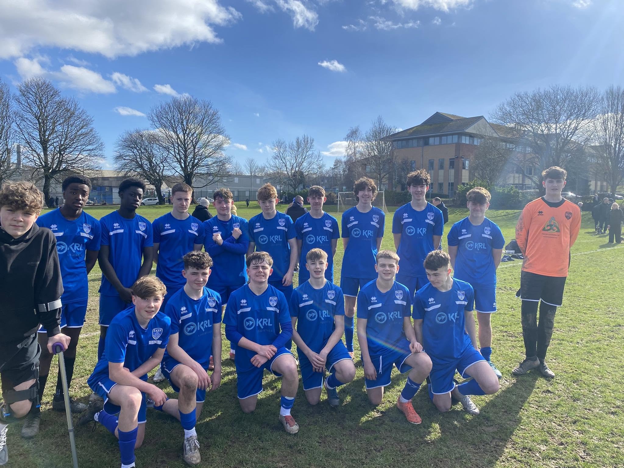 A group of young male soccer players in blue uniform posing for a team photo on a grassy field, with a few people and buildings in the background under a partly cloudy sky.