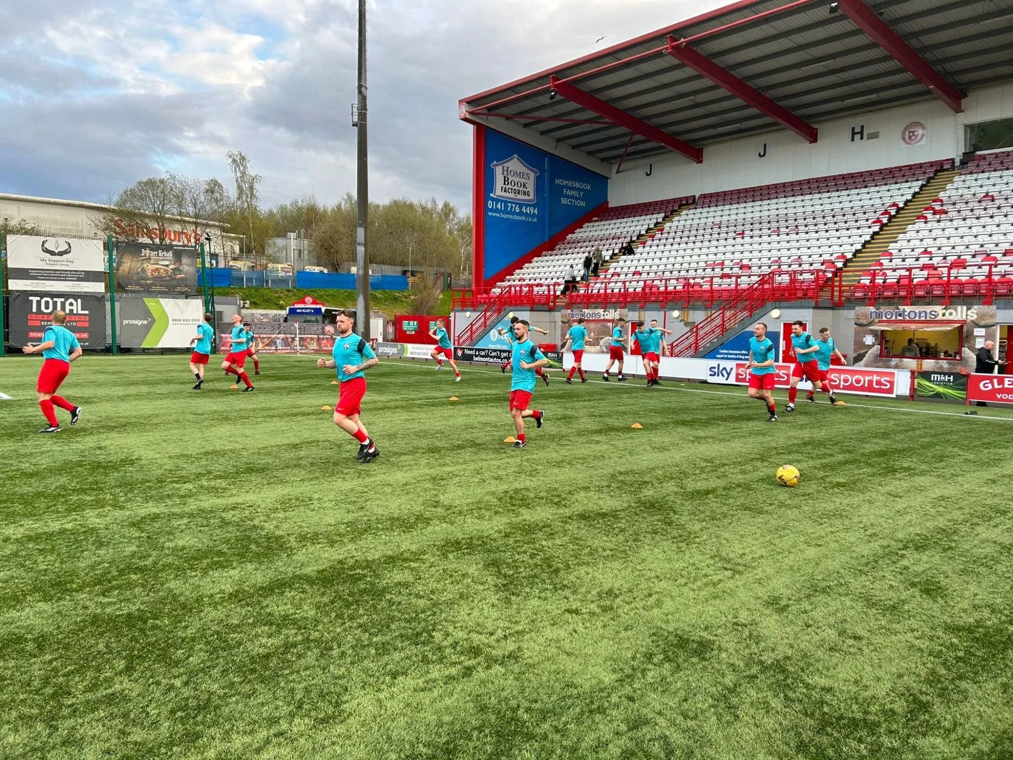 A group of soccer players in blue jerseys and red shorts practicing on a green grass field in a stadium. Some players are running and others are standing near small orange cones. The stadium has a large red and white seating area in the background and advertisements around the field.