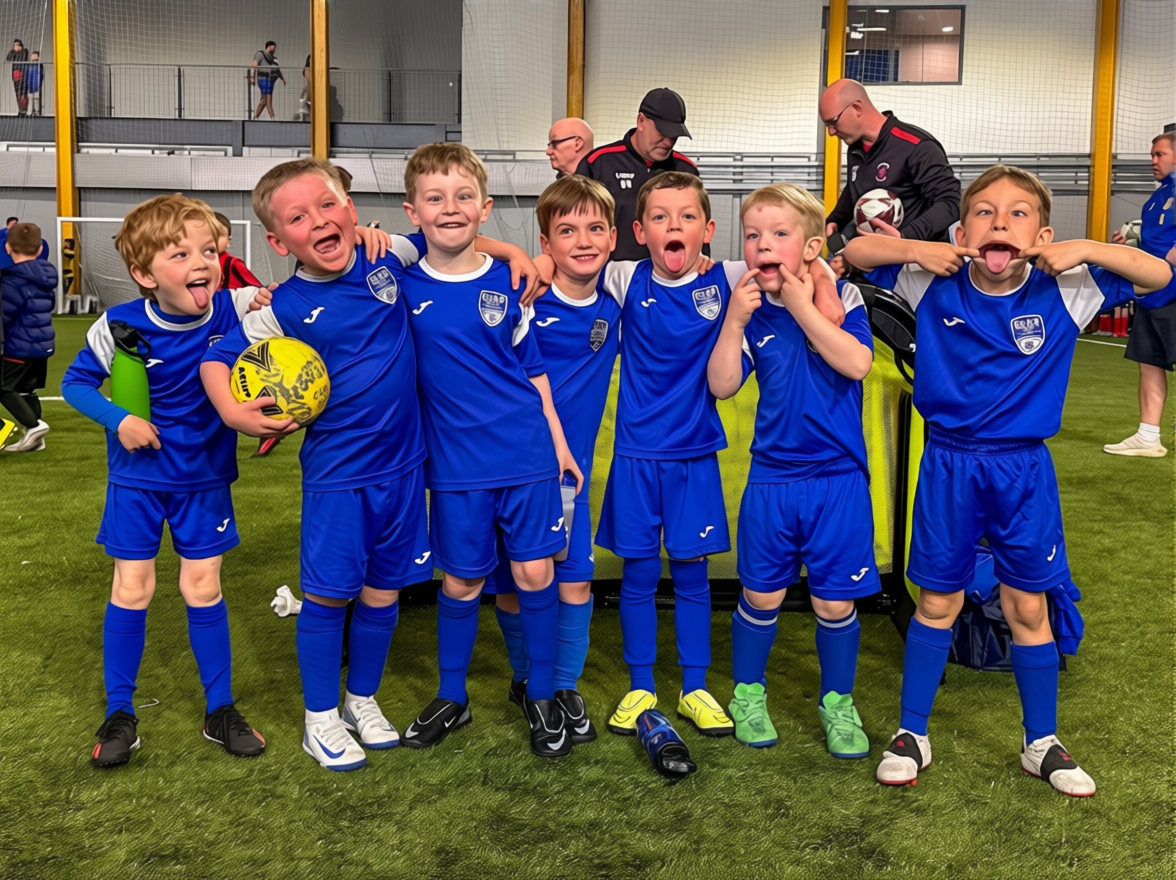 A group of young boys in blue soccer uniforms posing for a photo inside an indoor sports facility, with two coaches or adults standing behind them.