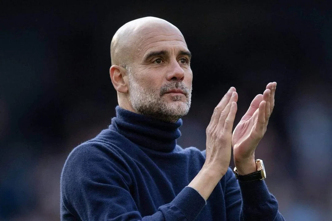 A man with a shaved head and gray beard wearing a dark blue turtleneck sweater, looking solemn and holding his hands up.