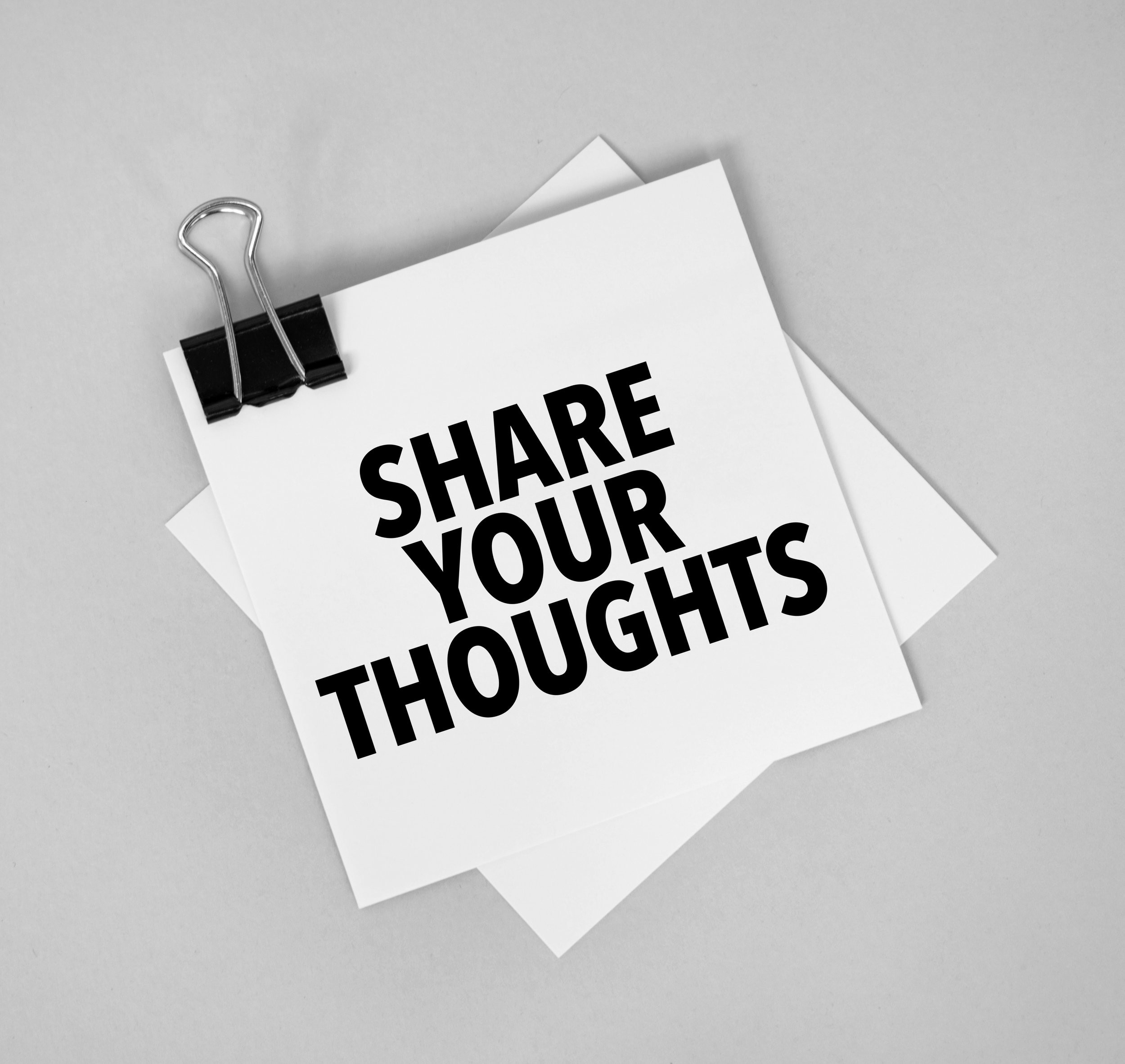 A black binder clip holding a white piece of paper with bold text that says 'SHARE YOUR THOUGHTS' against a light gray background.